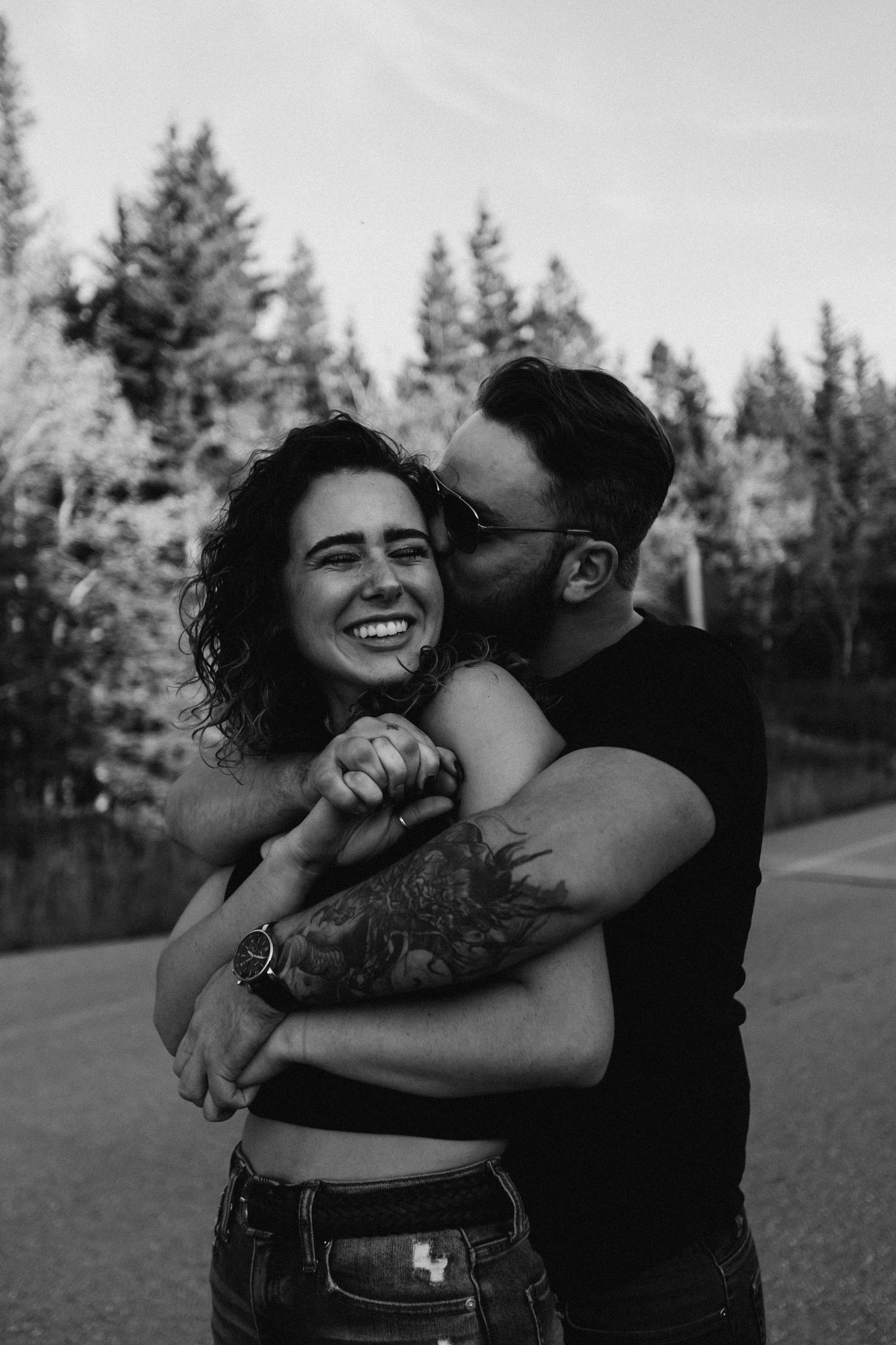 Black and white photo of a laughing woman being hugged and kissed by her partner outdoors.