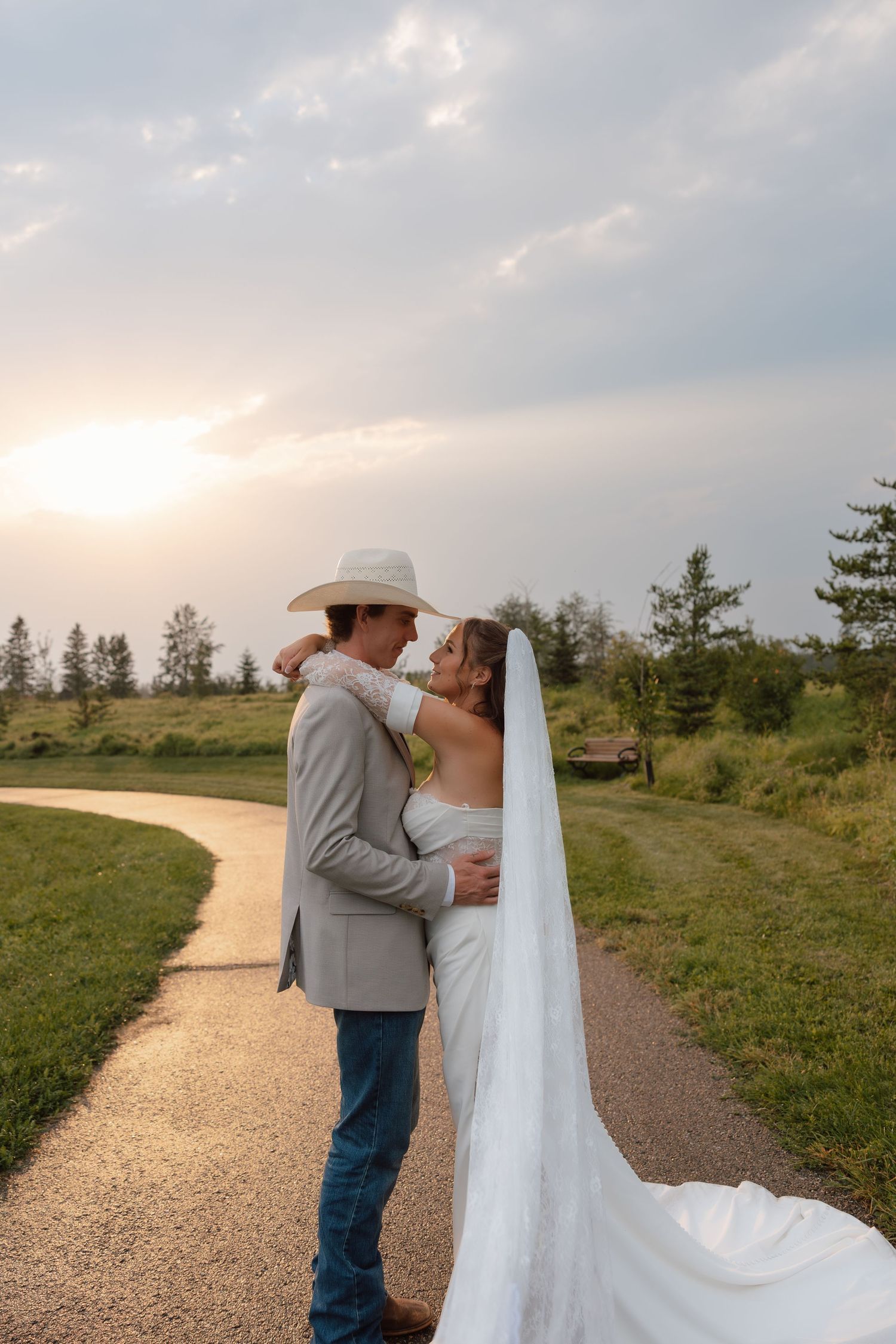 Bride and groom embrace on a winding path at sunset, she in a flowing white gown with veil, he in gray suit and cowboy hat.