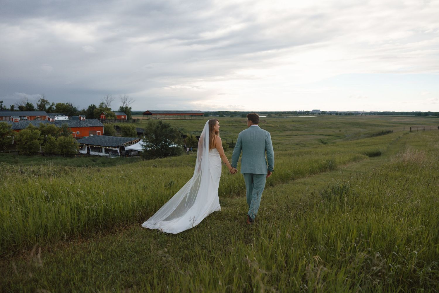 Bride and groom walking hand-in-hand through lush green fields at dusk, bride wearing flowing white gown with long veil.
