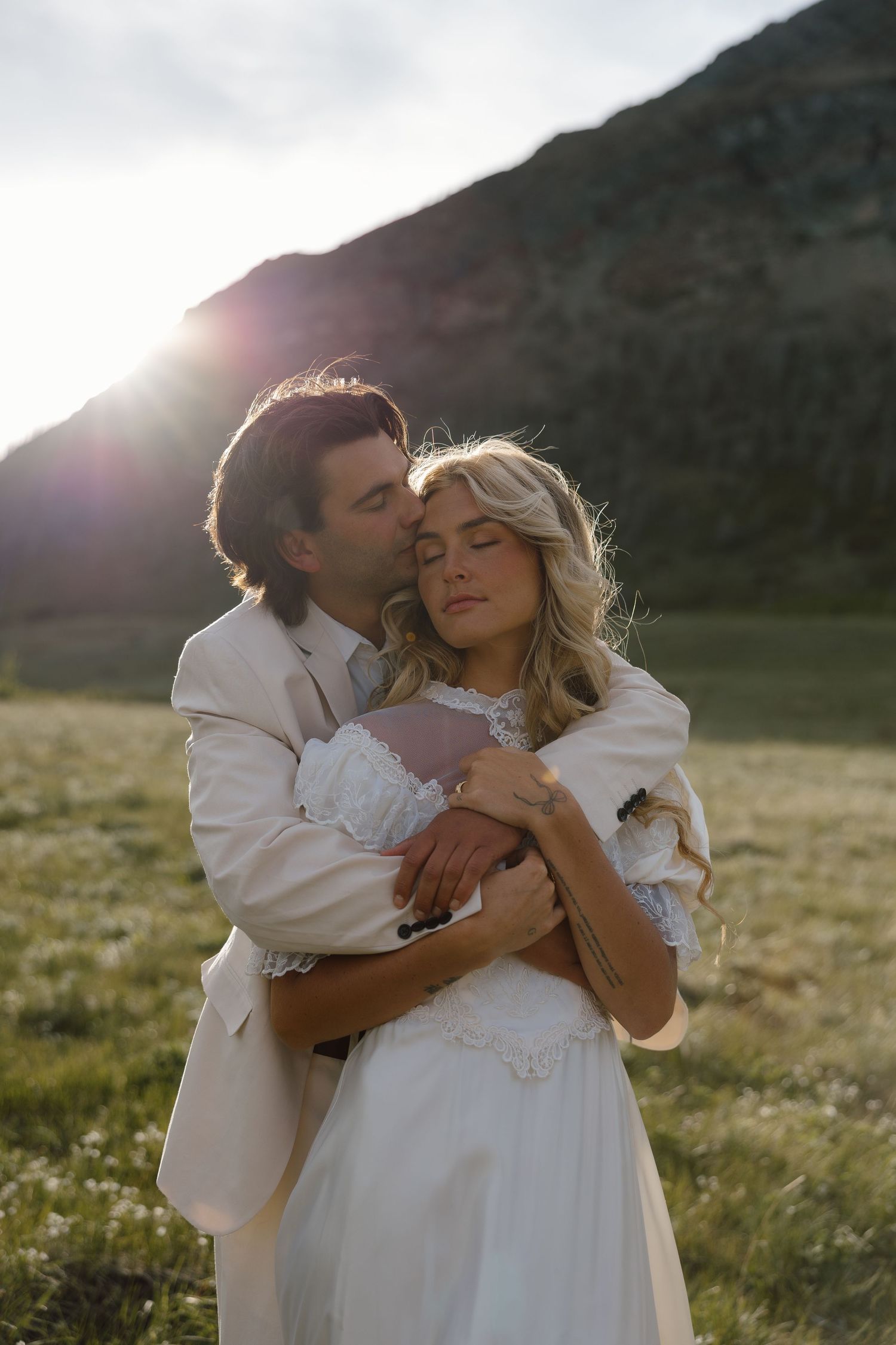 Couple embracing at golden hour in a mountain meadow, groom in tan suit kissing bride in white lace wedding dress.