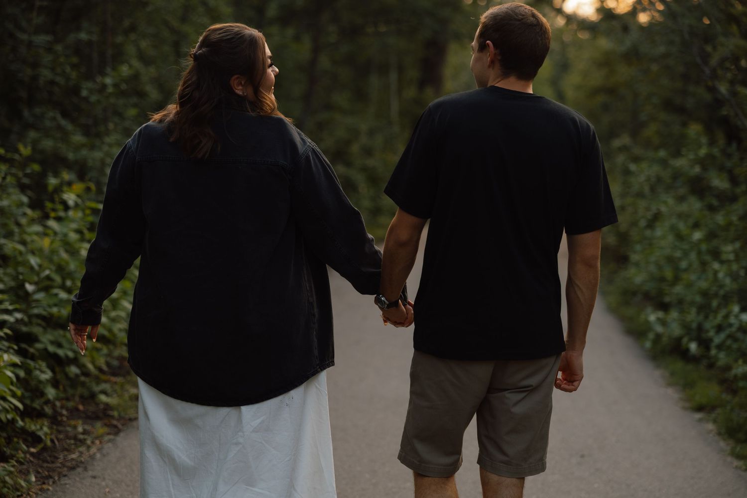 Couple holding hands walking on a tree-lined path, both wearing black tops, viewed from behind.