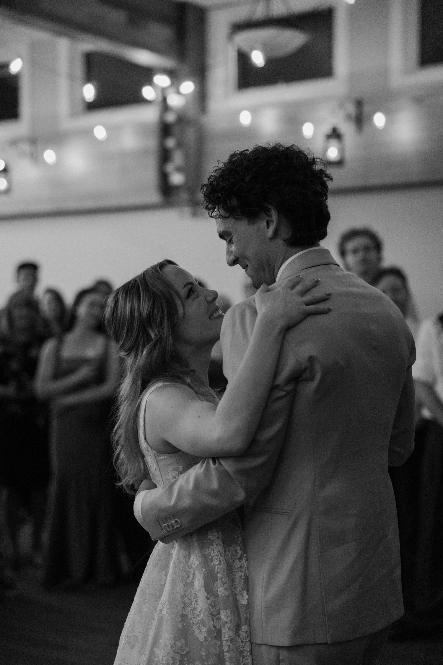 Black and white photo of a couple sharing their first dance at a wedding reception with guests watching in the background.