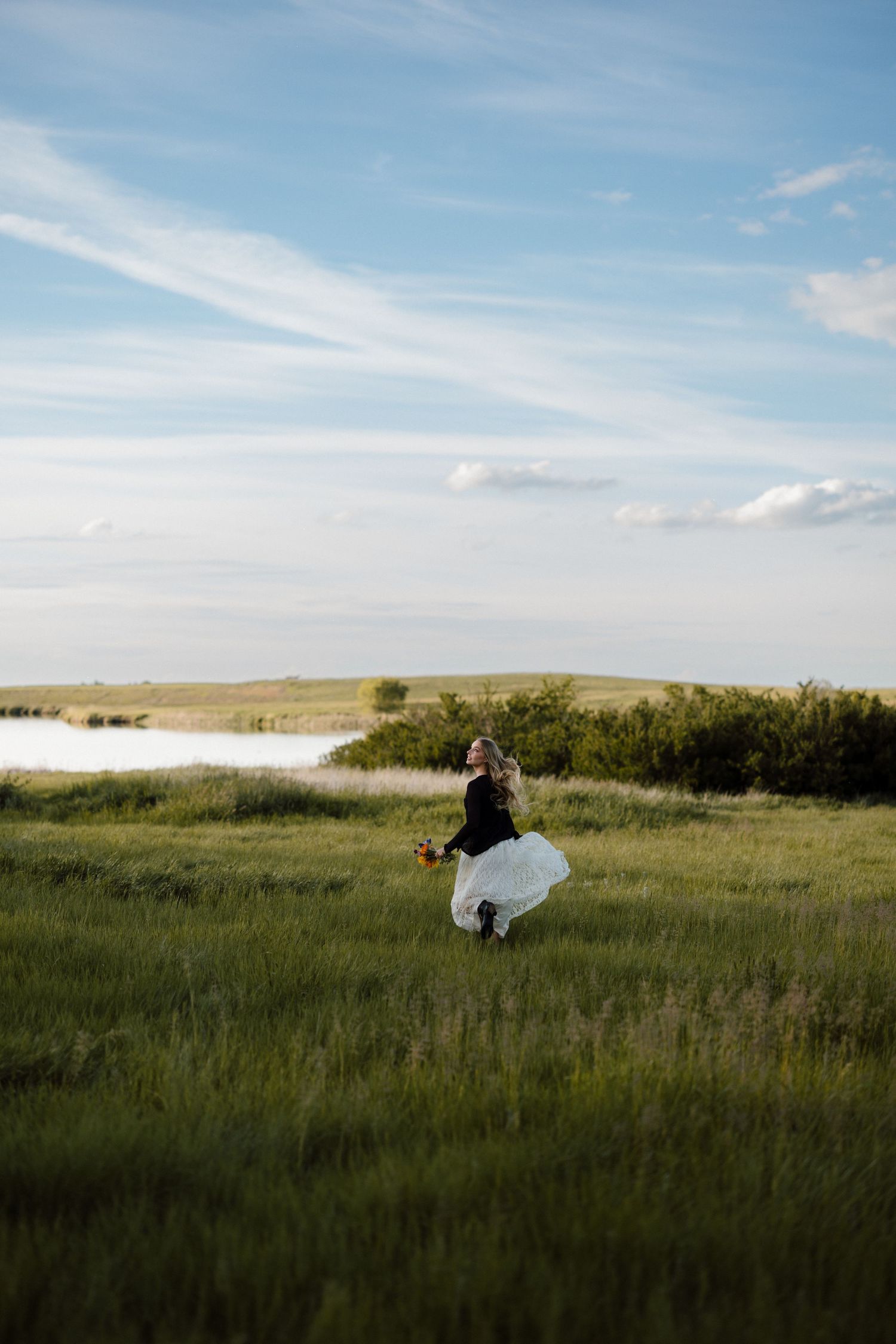A bride in a white gown twirls joyfully in a lush green field with a scenic lake backdrop.
