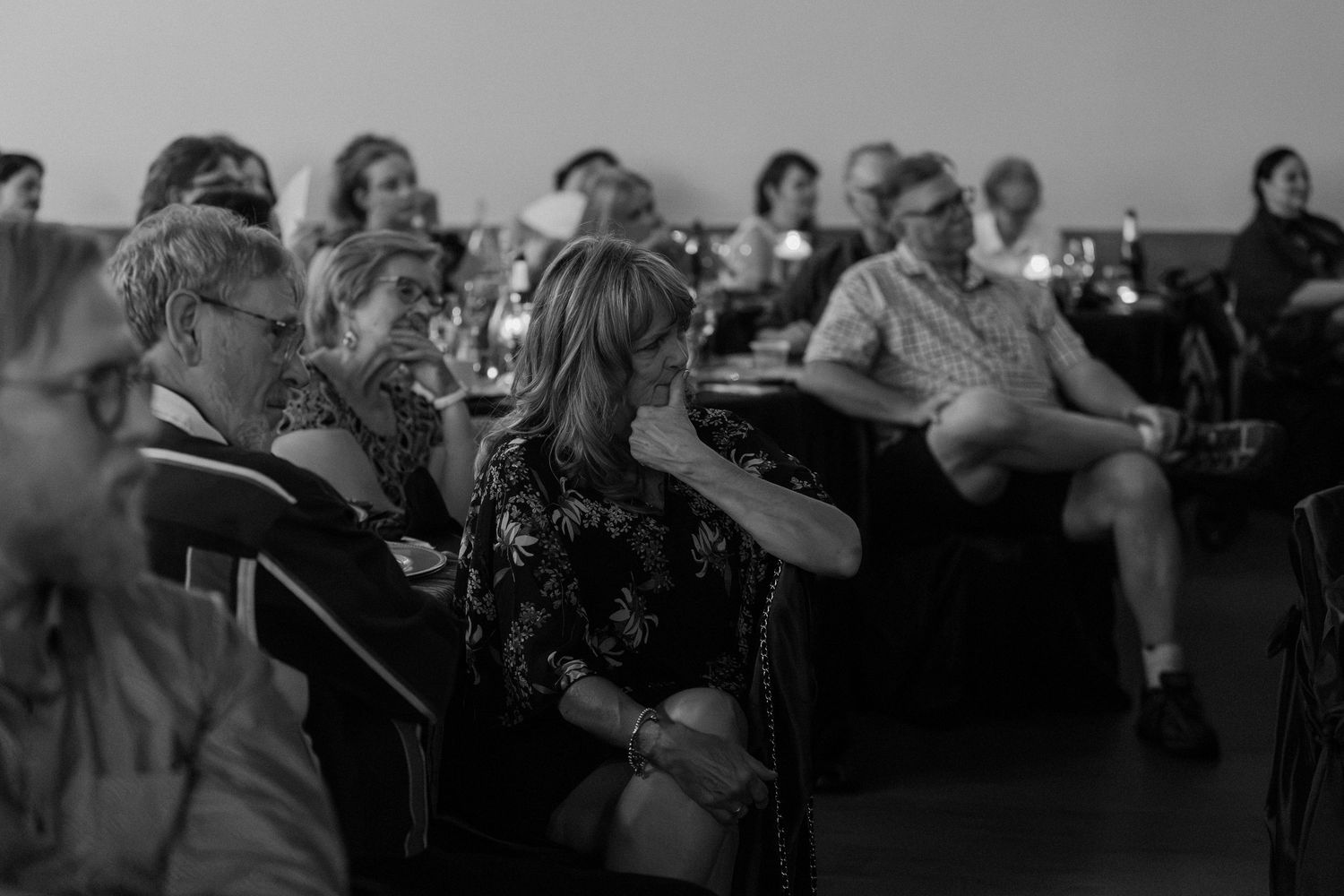 Black and white photo of an engaged audience seated at an indoor event, listening attentively in a dimly lit venue.