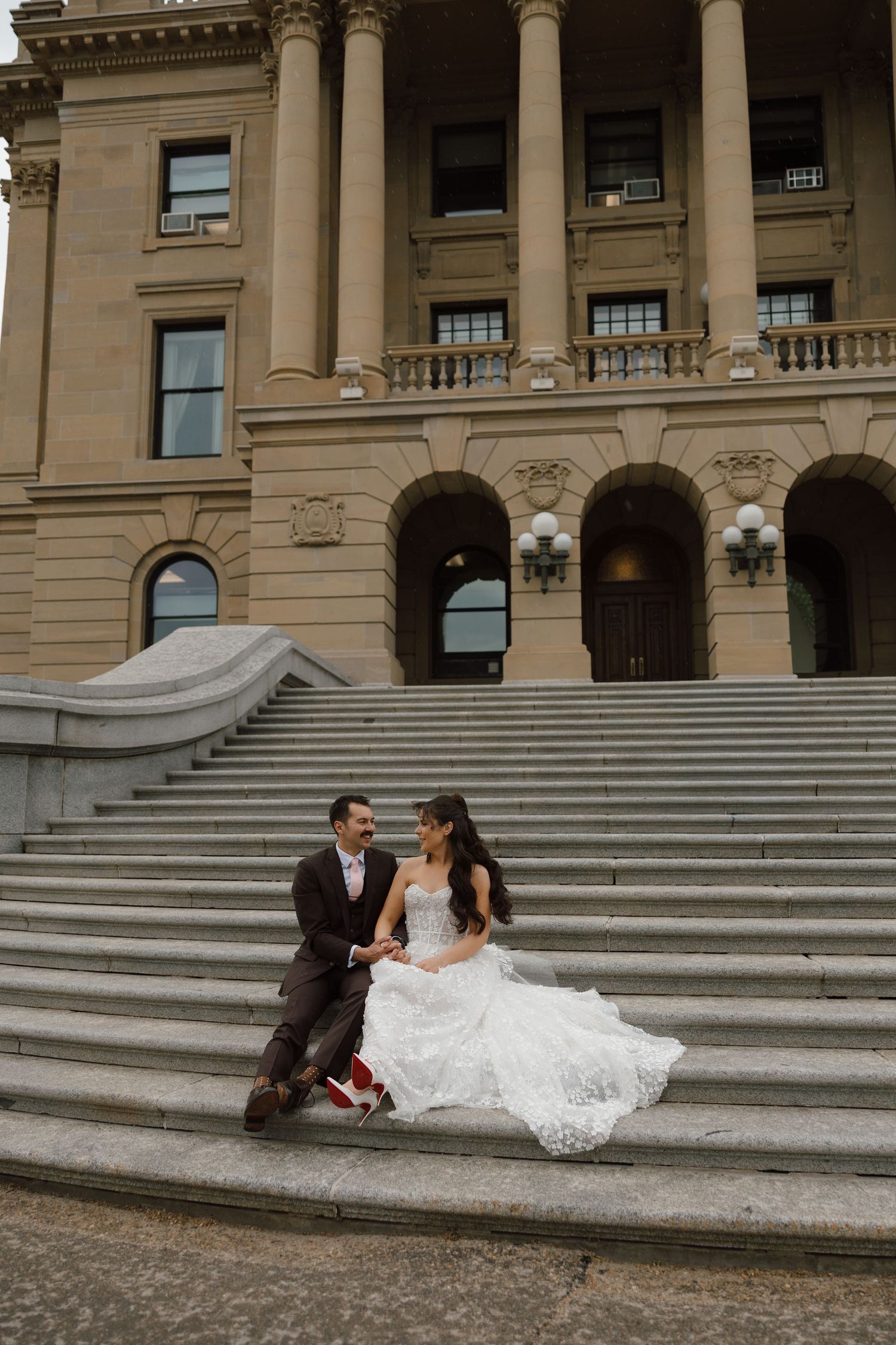 Bride and groom sitting on grand stone steps of a classical building, sharing a romantic moment in wedding attire.