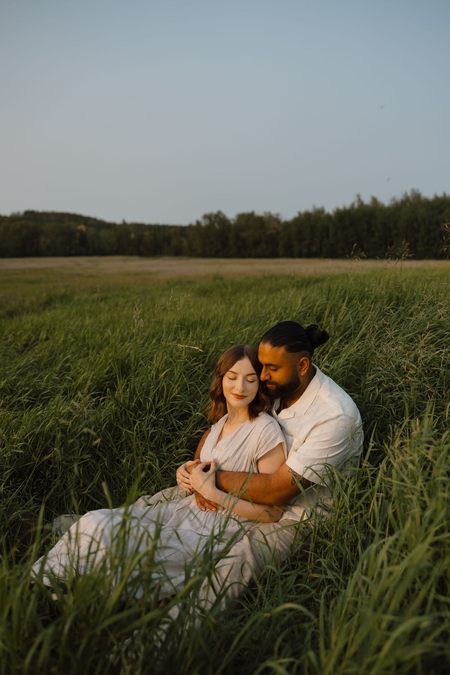 Couple sitting in tall green grass field at golden hour, man embracing pregnant woman from behind in romantic outdoor setting.