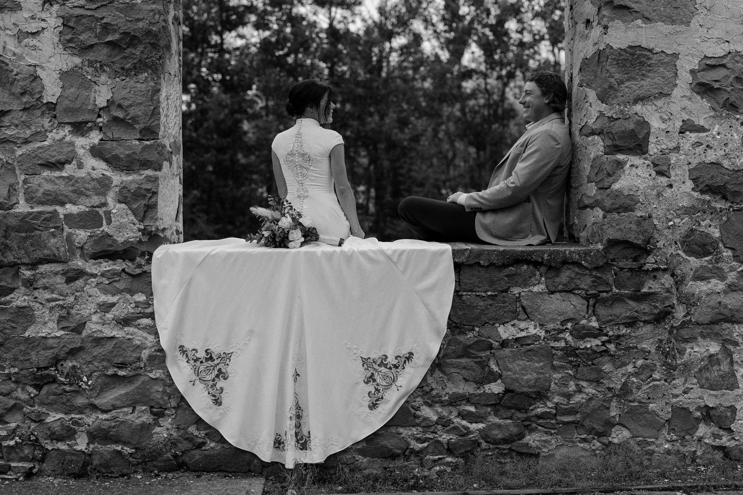 Black and white photo of bride and groom at rustic stone wall window with floral tablecloth draped over ledge. Taken in Alberta, Canada.