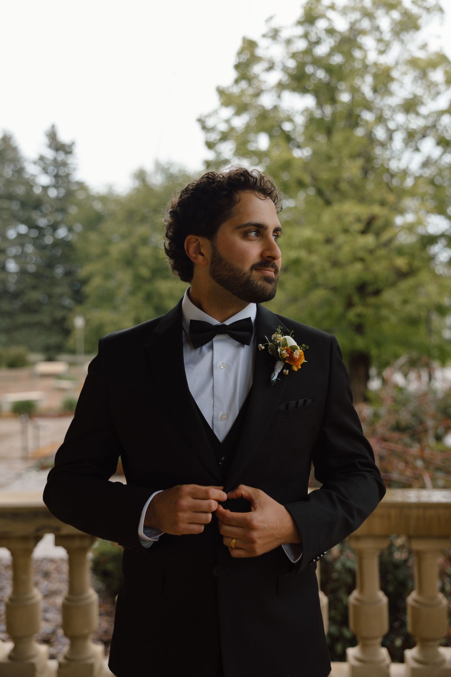 Dapper groom in black tuxedo with bow tie and floral boutonniere stands on stone balcony surrounded by lush trees.