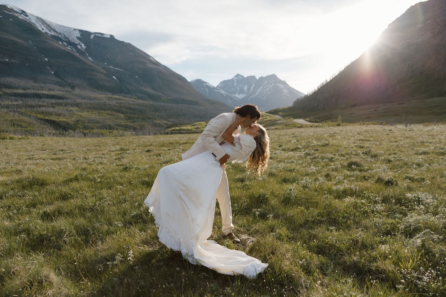 Couple sharing a romantic dip kiss in a lush green mountain meadow, bride in white gown, sun rays behind peaks.