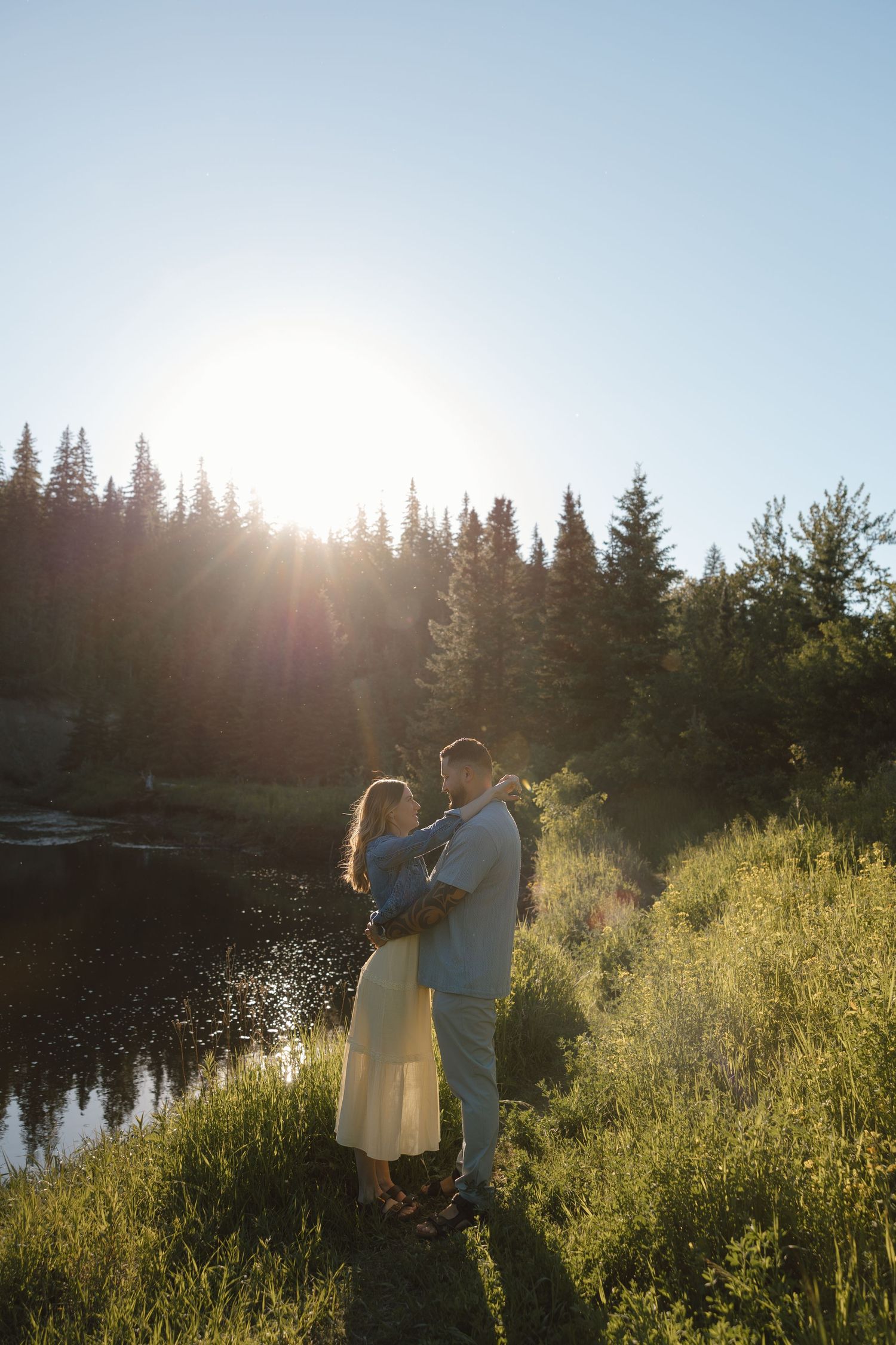 Couple embracing at golden hour by a forest lake, sun flaring behind evergreen trees, lush green meadow.