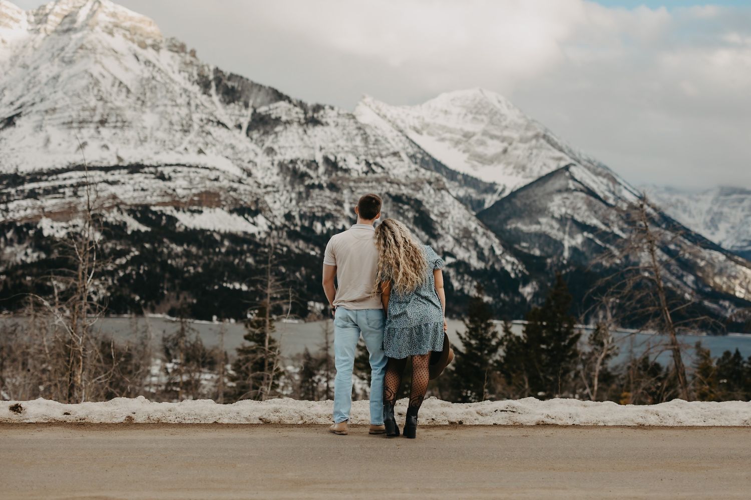 Couple standing arm in arm admiring snow-capped mountain peaks and pine forest landscape in winter. Taken in Waterton Lake National Park.