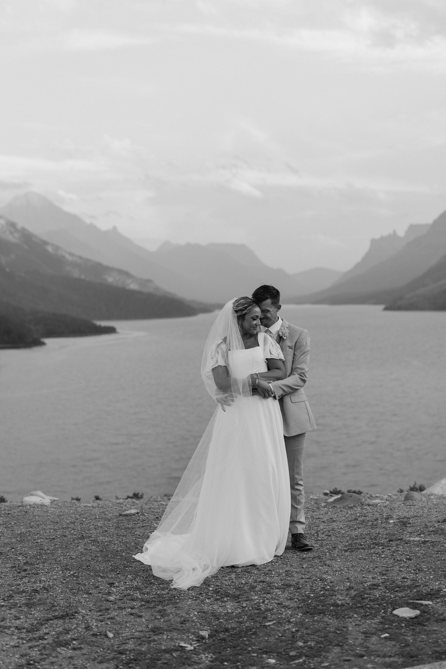 Bride and groom embrace on rocky lakeside with stunning mountain backdrop in black and white. Taken in Waterton National Park