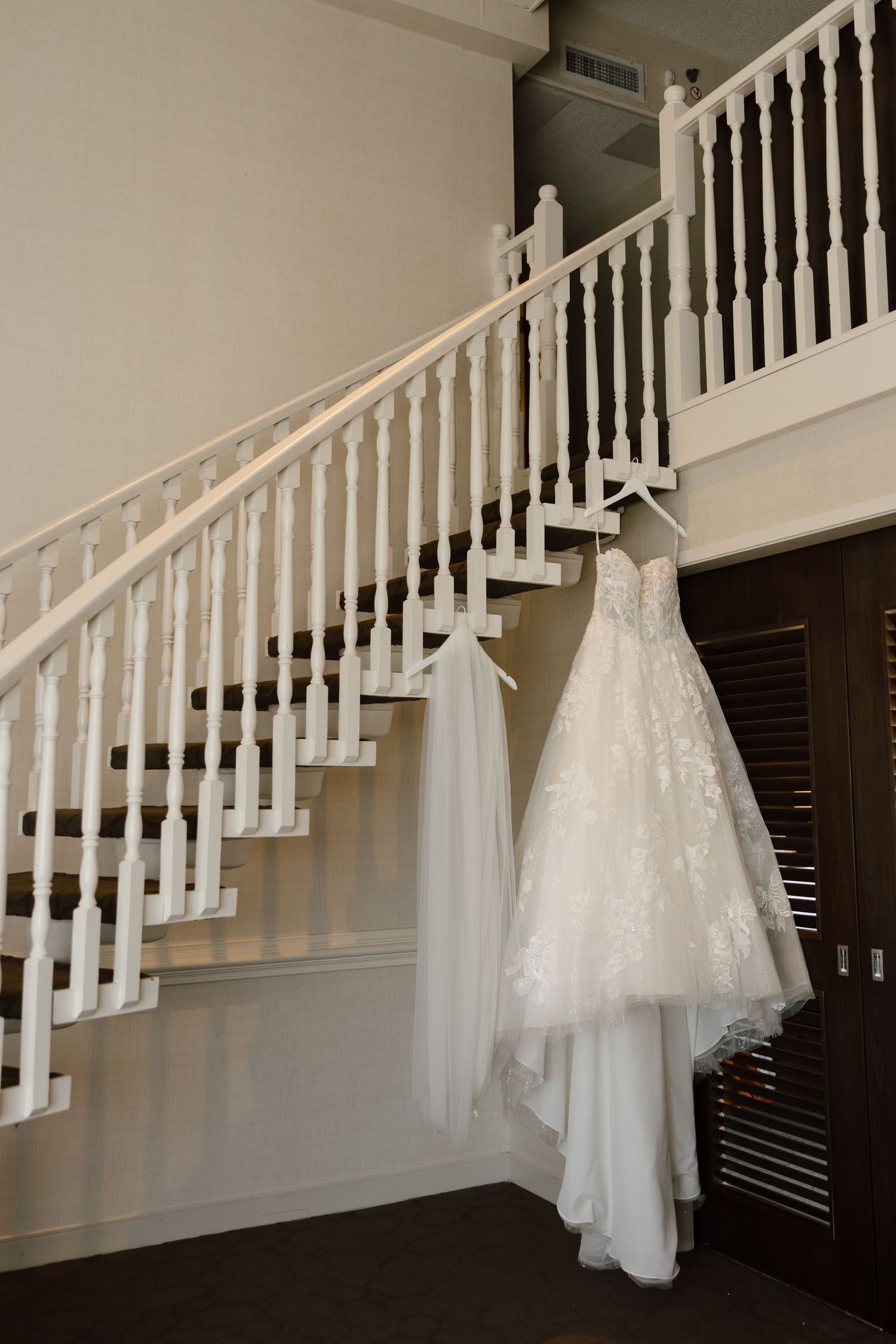 Elegant lace wedding dress hanging on white staircase railing in a bright, classic venue foyer.