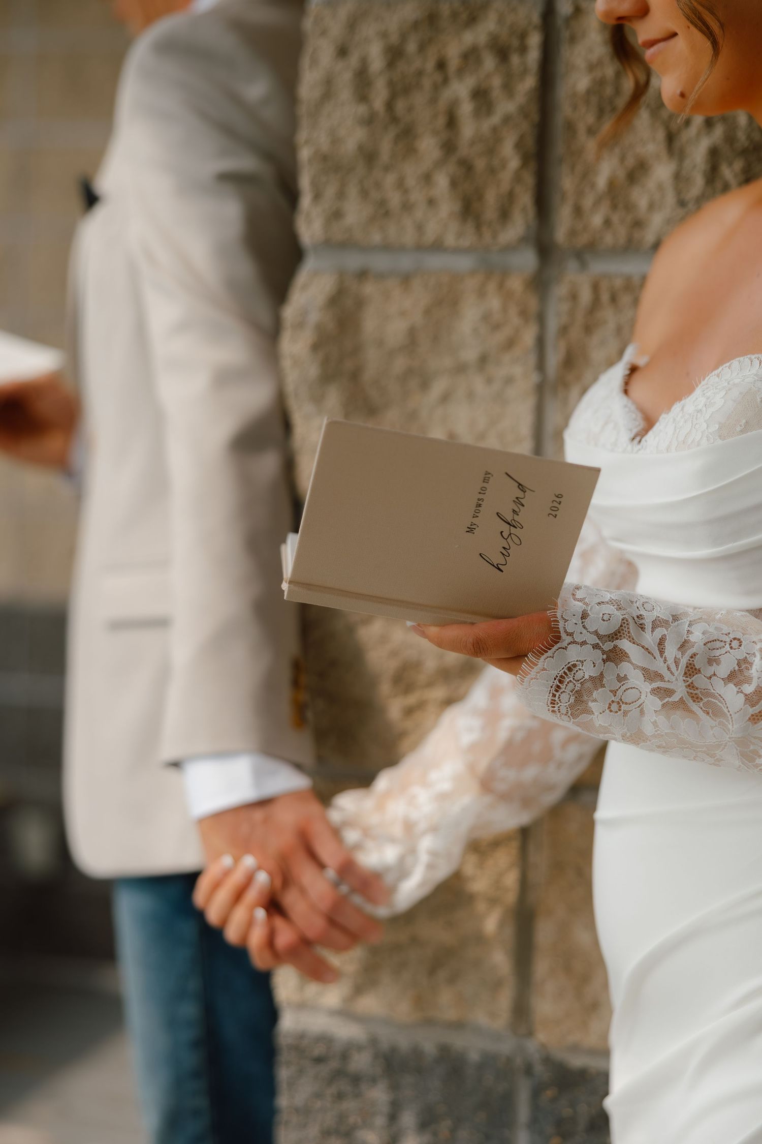 Bride in lace wedding dress holds vows booklet and groom's hand during outdoor wedding ceremony.