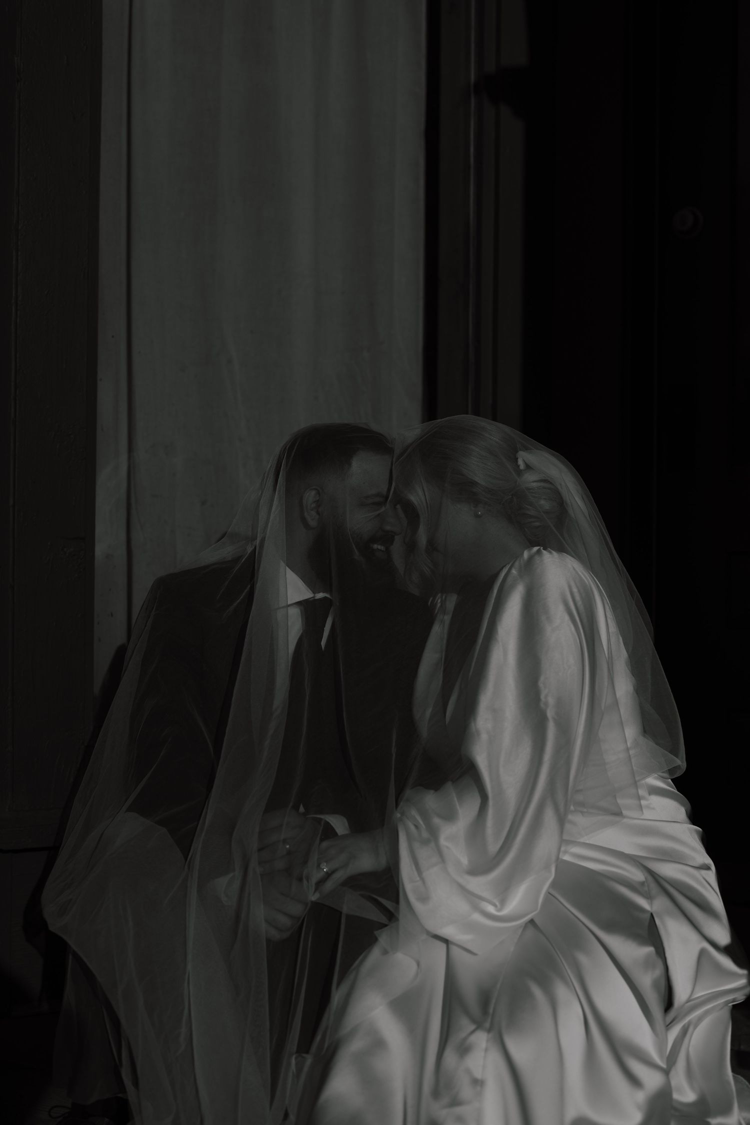 Dark romantic black and white portrait of bride and groom laughing together under a veil.