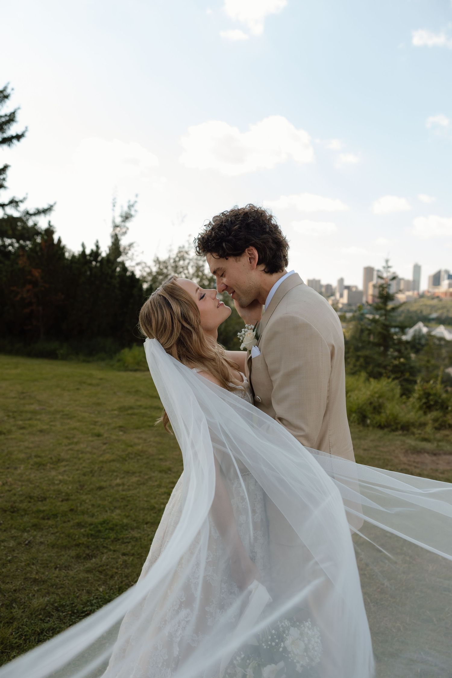 Bride and groom nearly kissing outdoors, her veil flowing in wind, city skyline and trees in background.