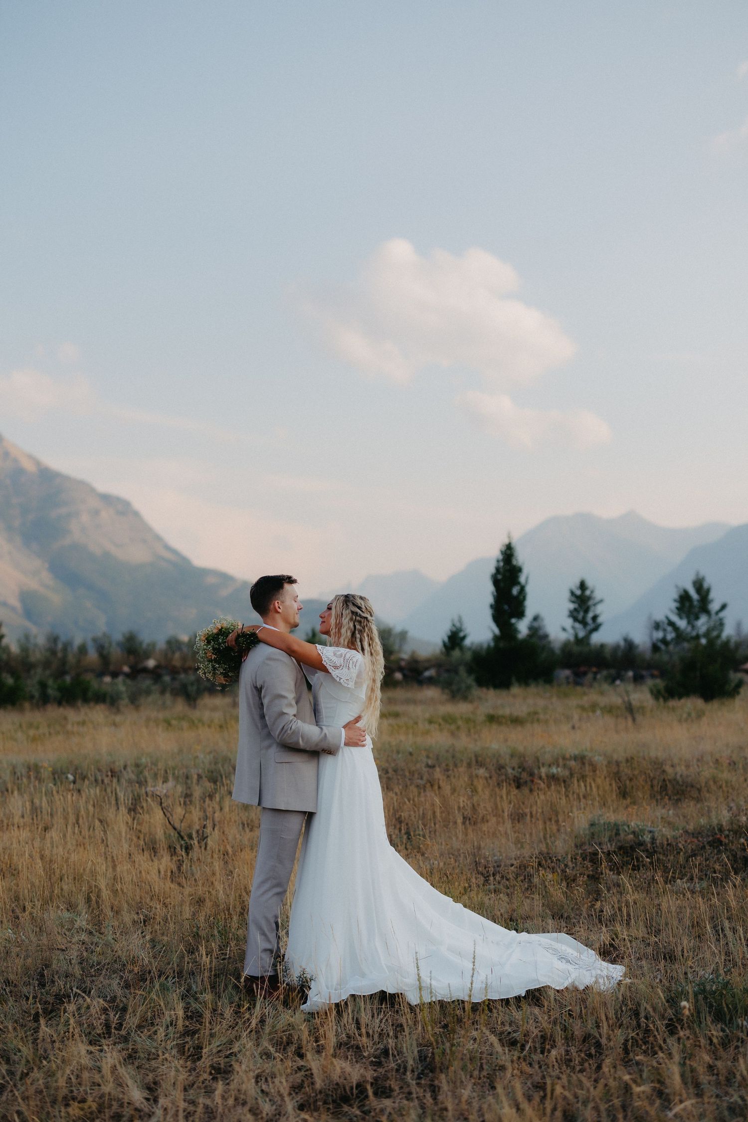 Bride and groom embracing in golden field with dramatic mountain range and soft sky in background. Taken in Waterton National Park.