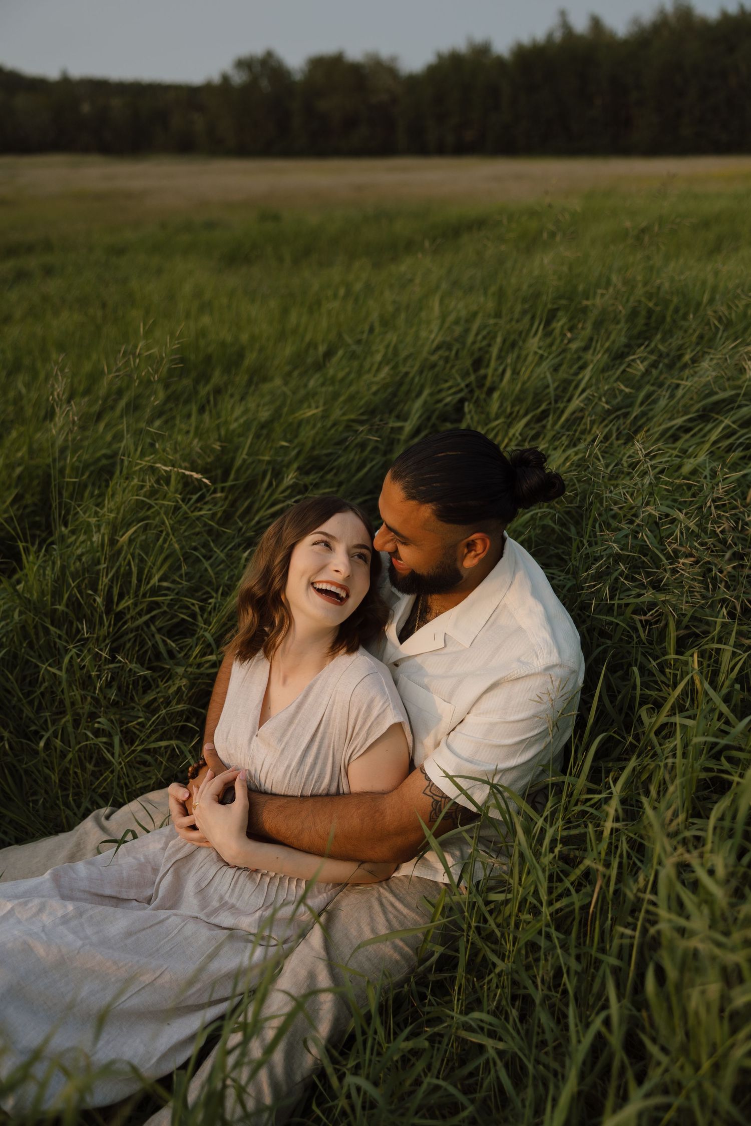 A couple laughing and embracing in a lush green field at golden hour, woman in white dress leaning against man in white shirt.