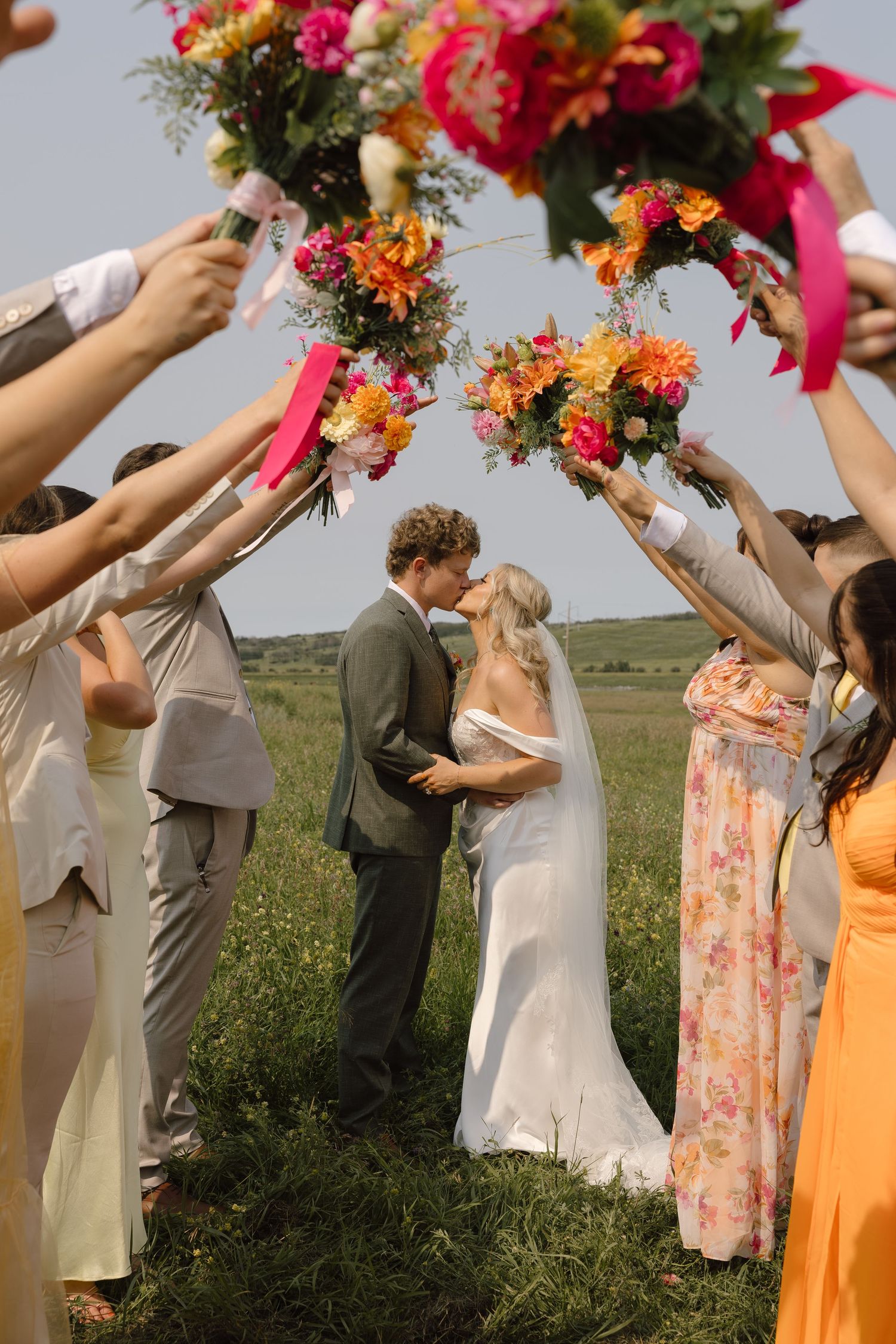 Bride and groom share first kiss under arch of colorful bouquets held by wedding party in green field.