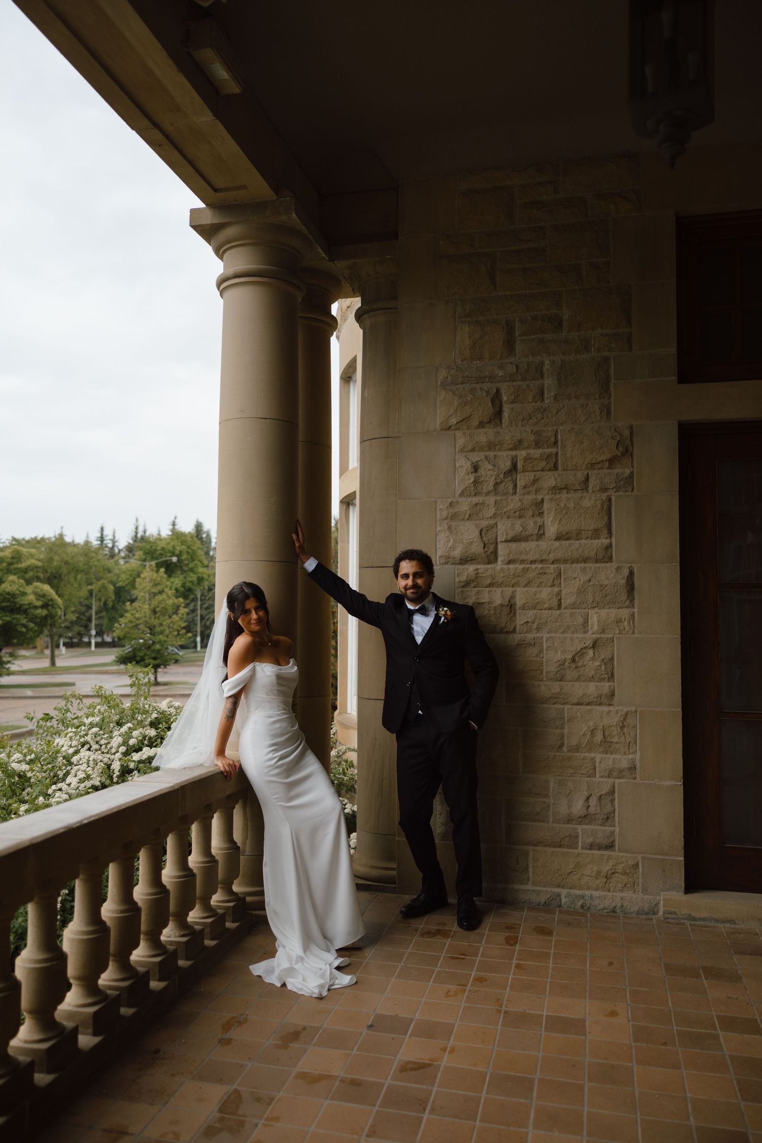 Bride in white gown and groom in black tuxedo posing on ornate stone balcony with columns on their wedding day.