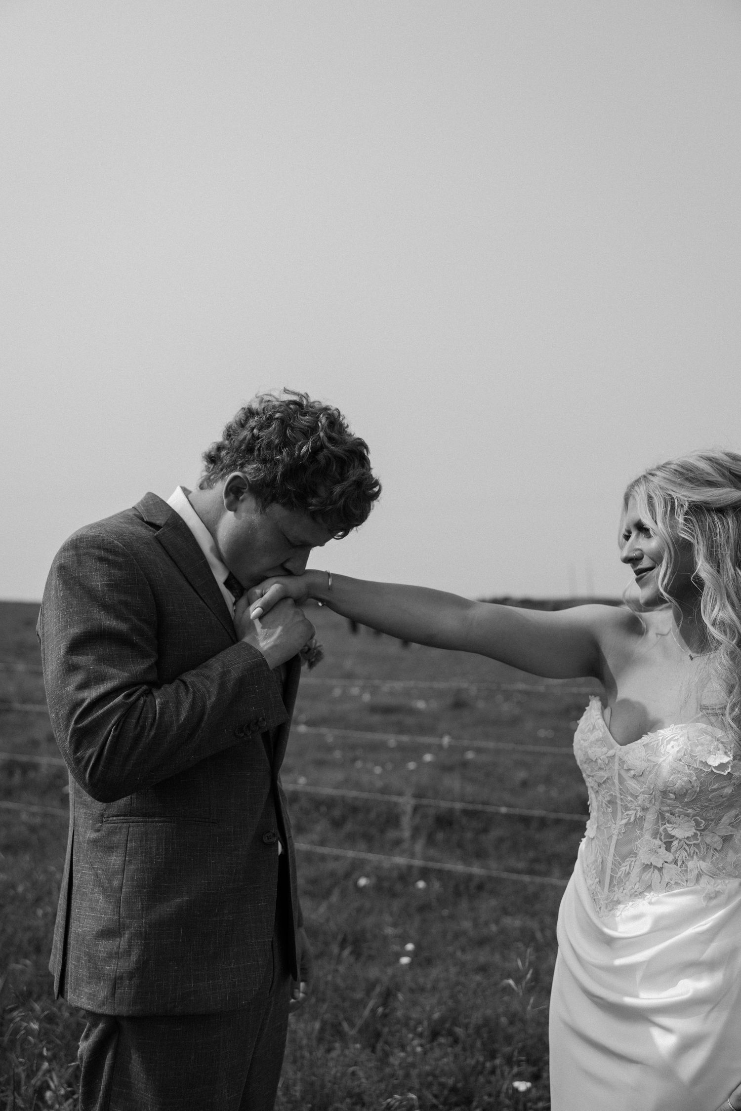 Black and white photo of a groom kissing his bride's hand outdoors in a field on their wedding day.