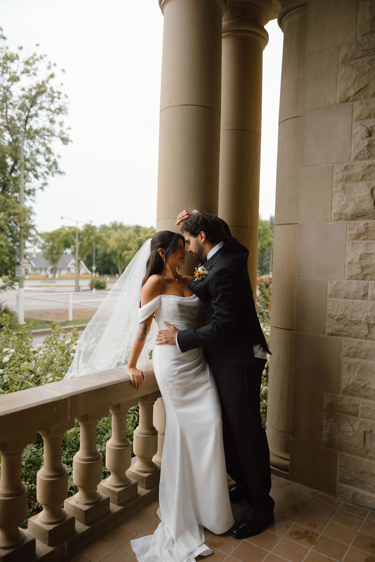 Bride in white gown and groom in black suit share a romantic moment on a stone balcony with columns.