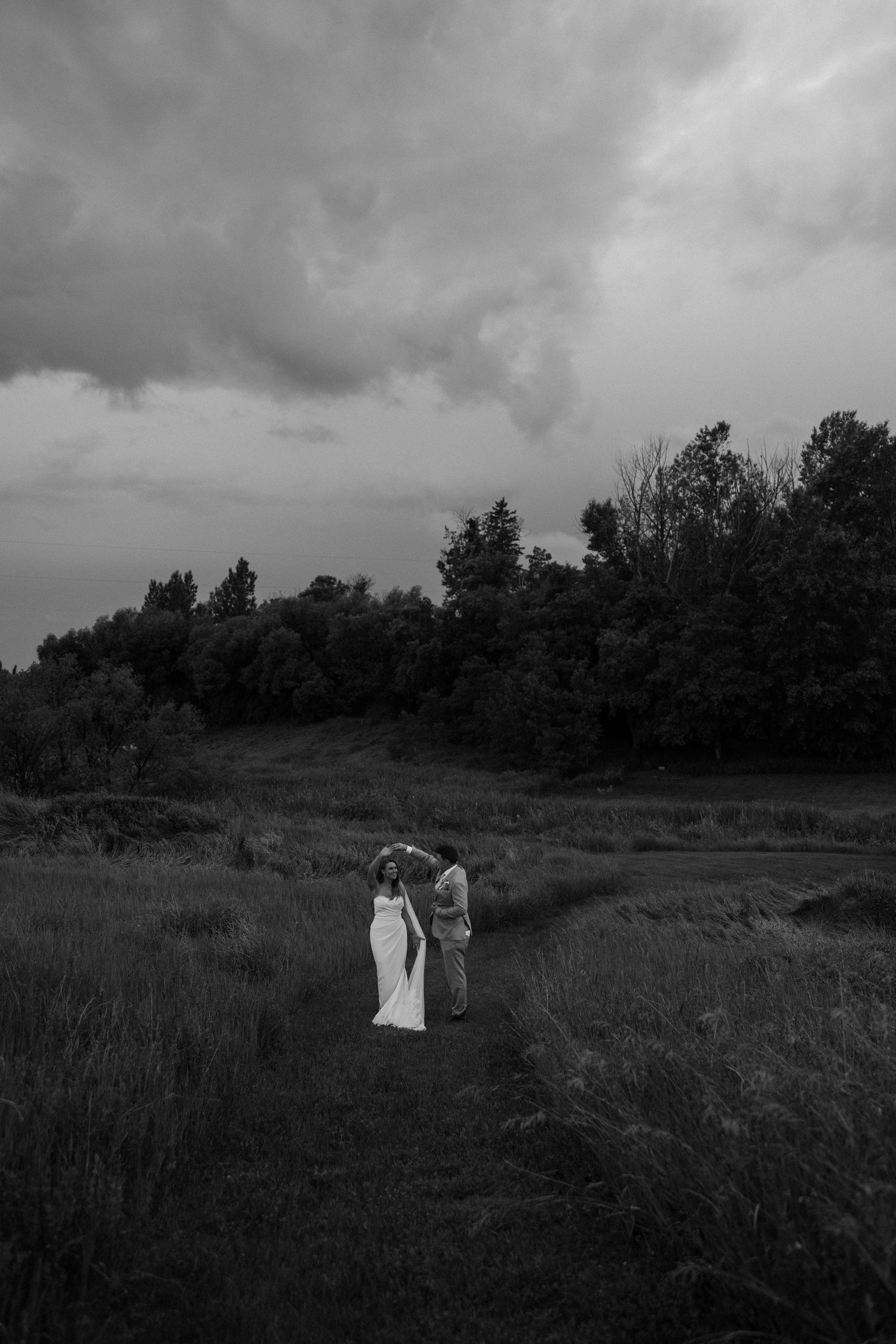 Black and white photo of a bride and groom walking through a meadow path with dramatic stormy skies and trees.
