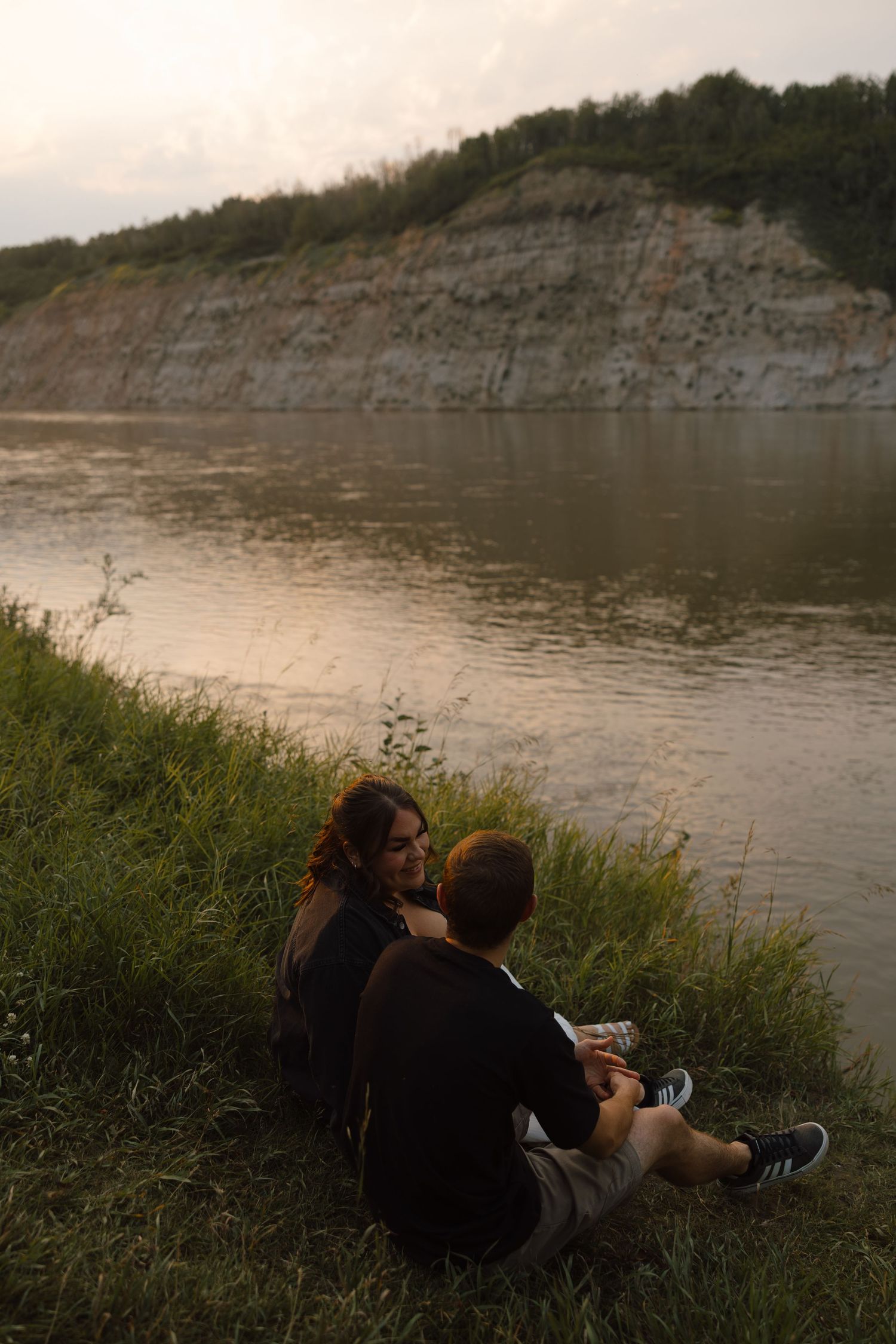 Couple sitting on grassy riverbank at dusk, with a rocky cliff and calm water in the background.