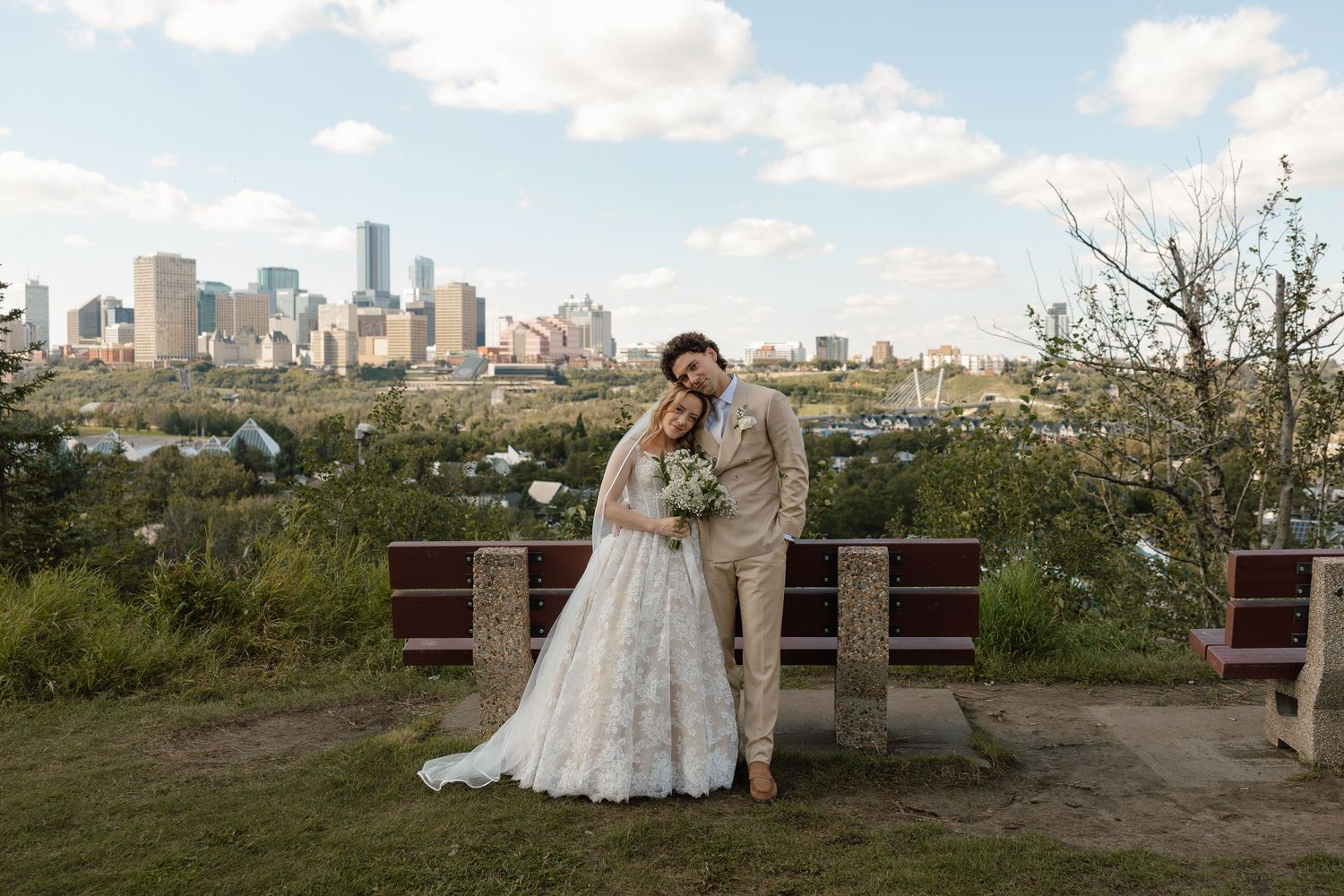 Bride and groom pose before Edmonton city skyline at outdoor wedding venue with lush greenery and cloudy sky.