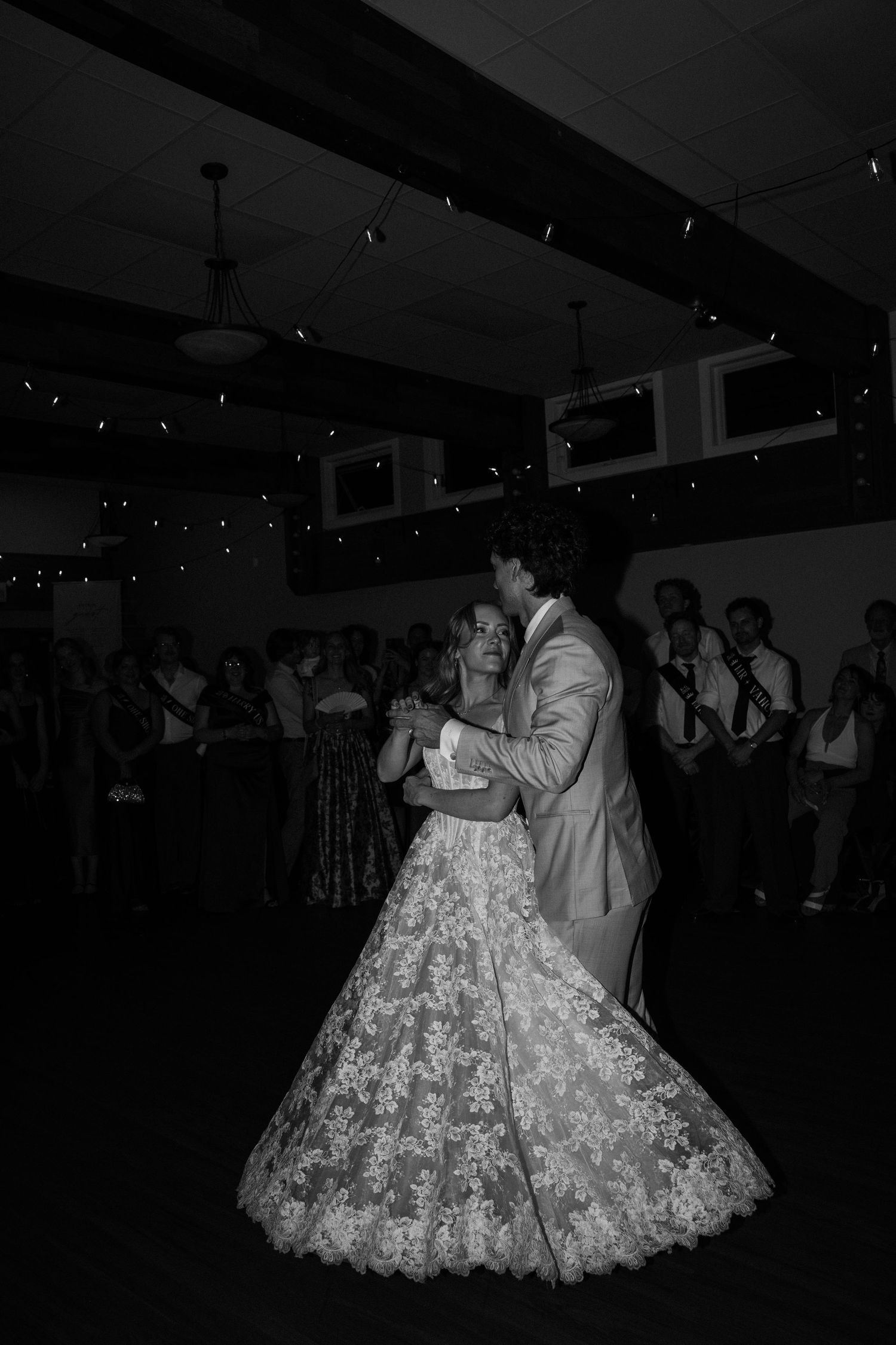 Black and white photo of a bride and groom sharing their first dance at a wedding reception with guests watching.