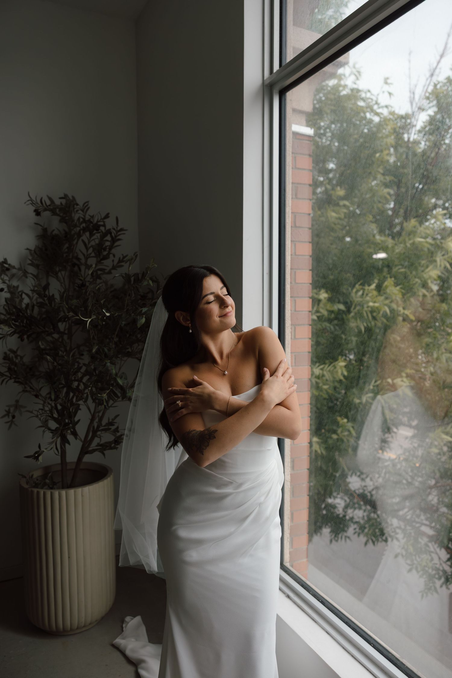 Bride in white gown and veil stands by large window, arms crossed, gazing outside with soft natural light.