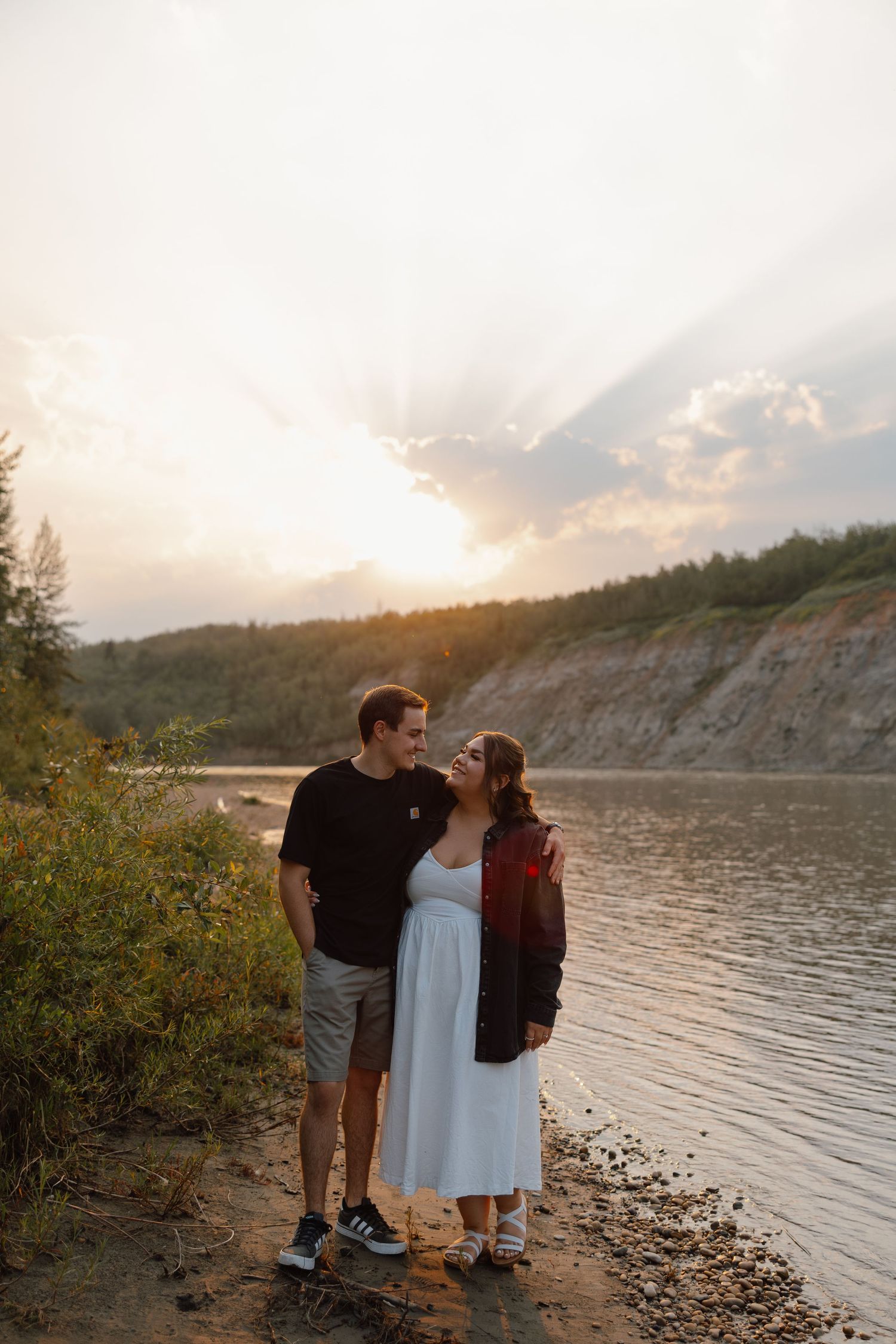 Couple embracing by a scenic lake at sunset, with golden rays breaking through clouds over a forested hillside.