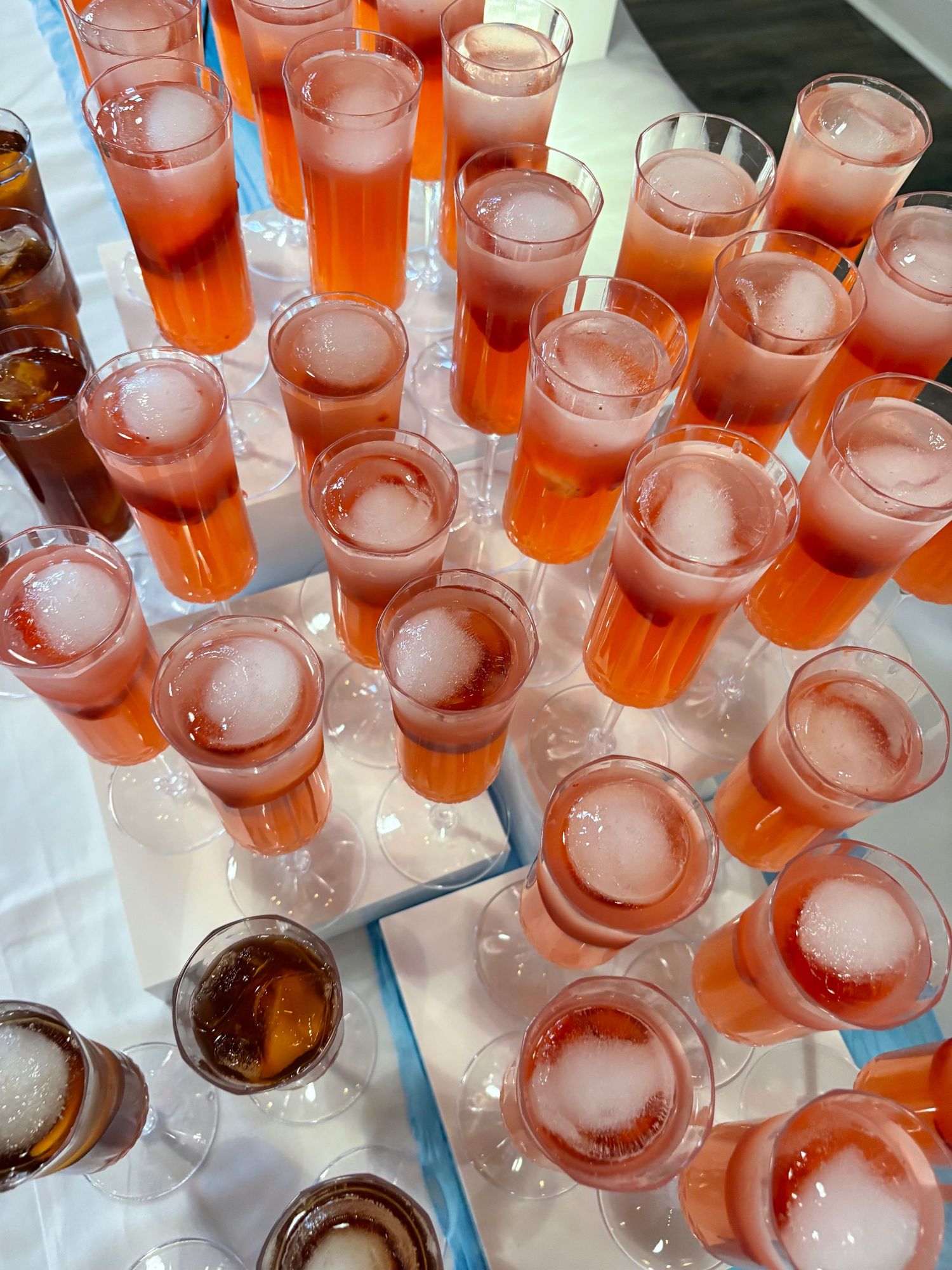 Rows of fizzing pink champagne flutes and brown cocktails arranged on a white table at a catered event.