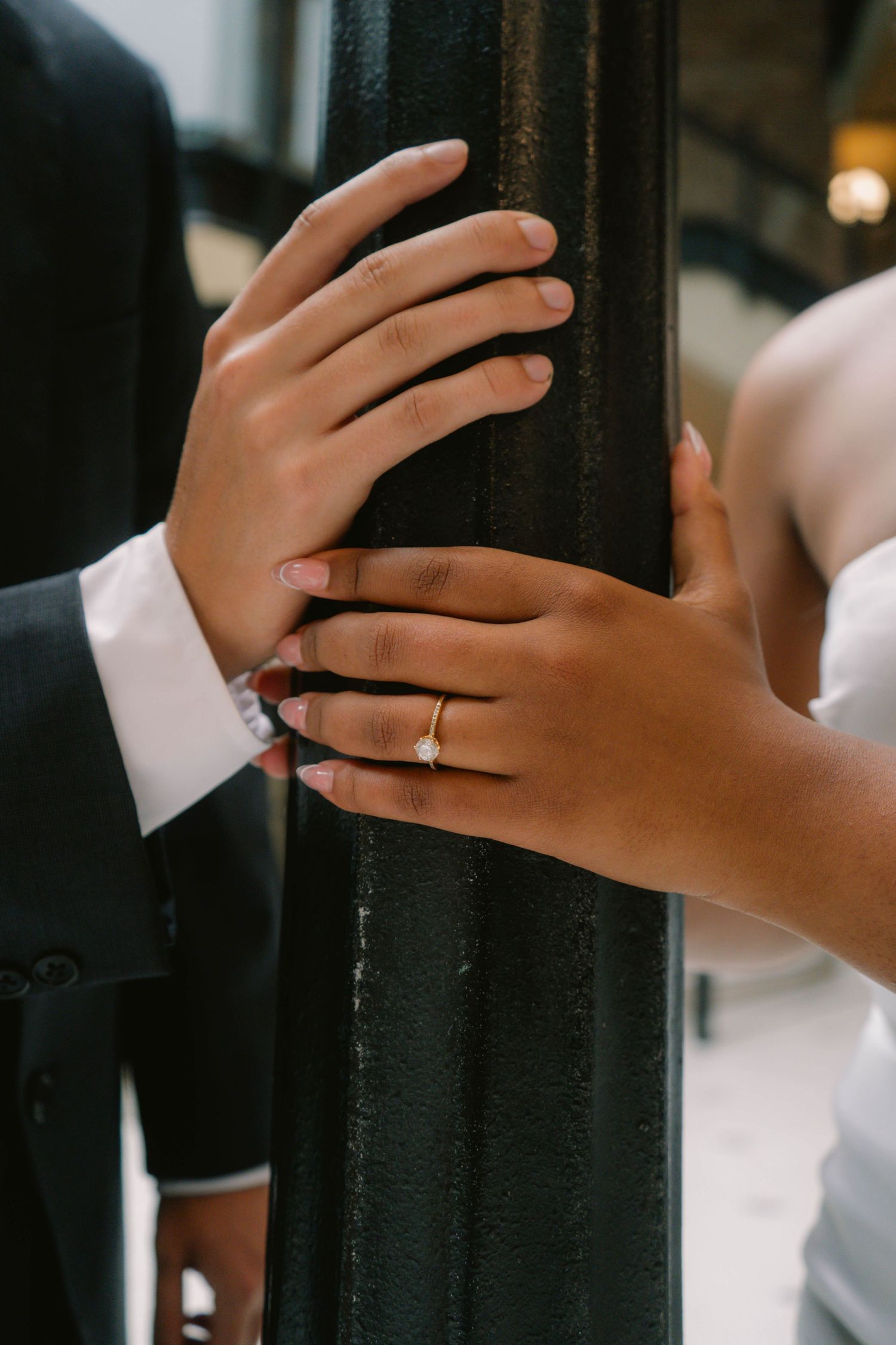 Close-up of bride and groom hands touching black iron post, bride wearing gold diamond engagement ring.