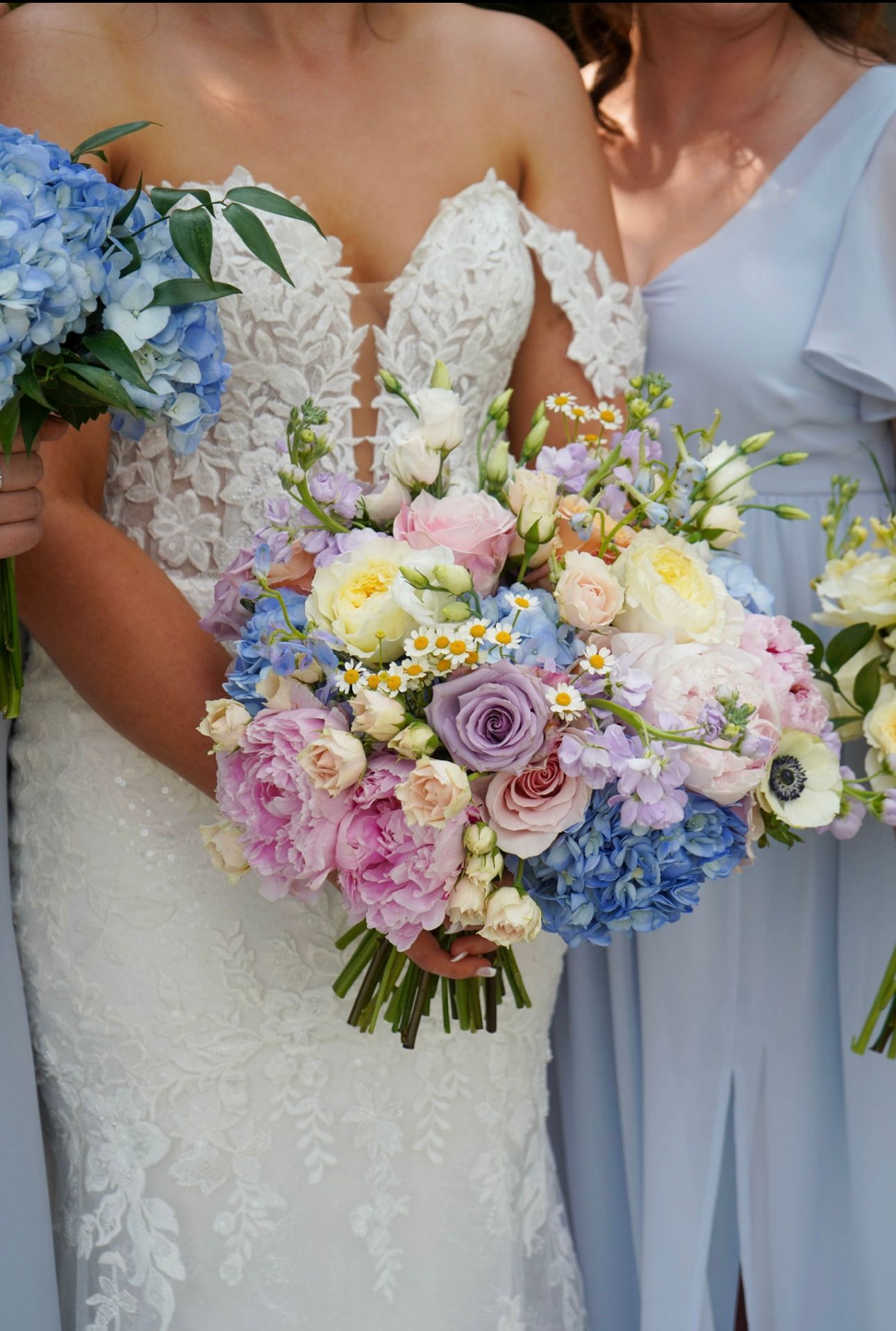 Bride in lace gown and bridesmaid holding pastel pink, purple, and blue floral bouquets.