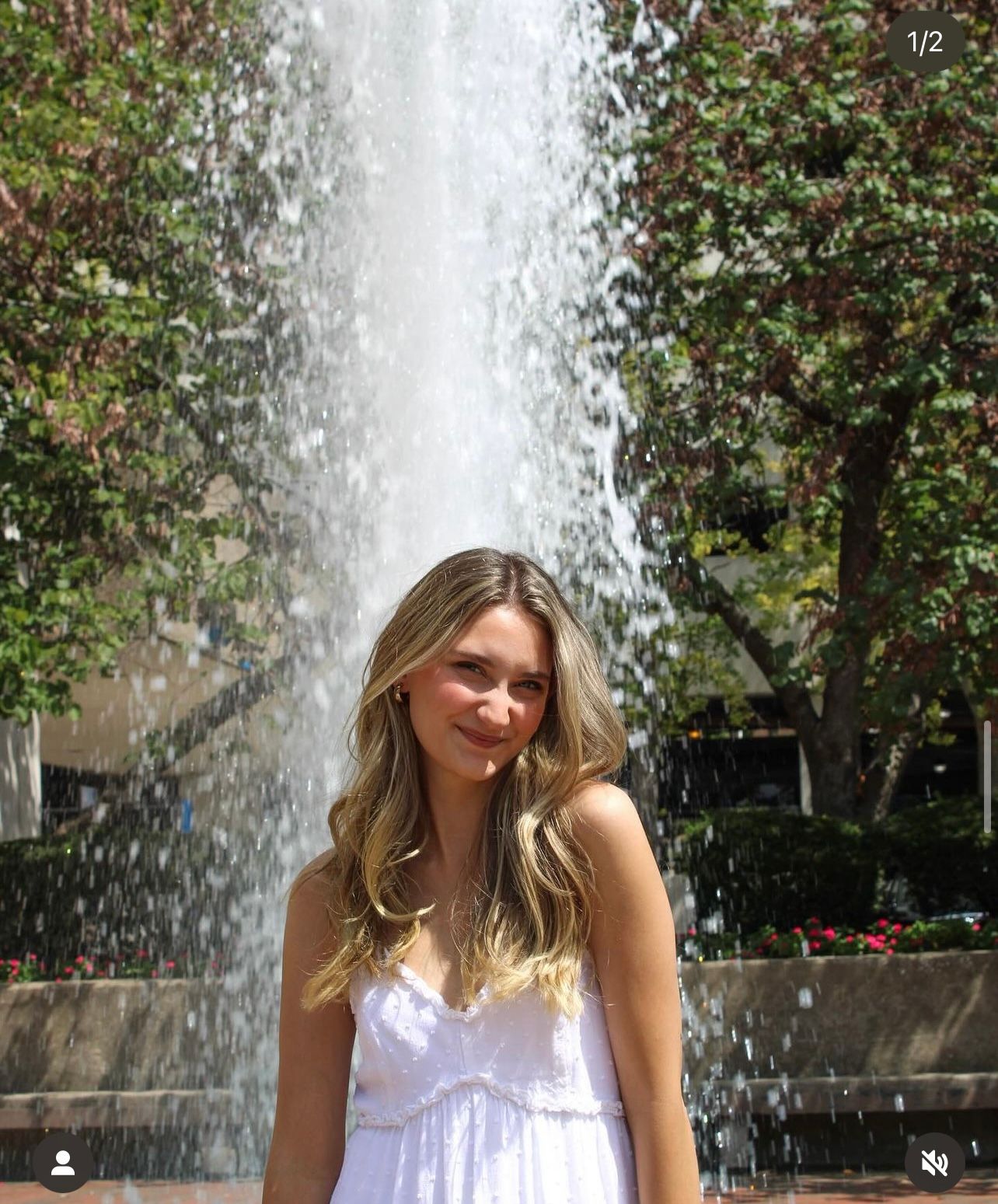 Young woman in white dress smiling in front of a large fountain outdoors on a sunny day.