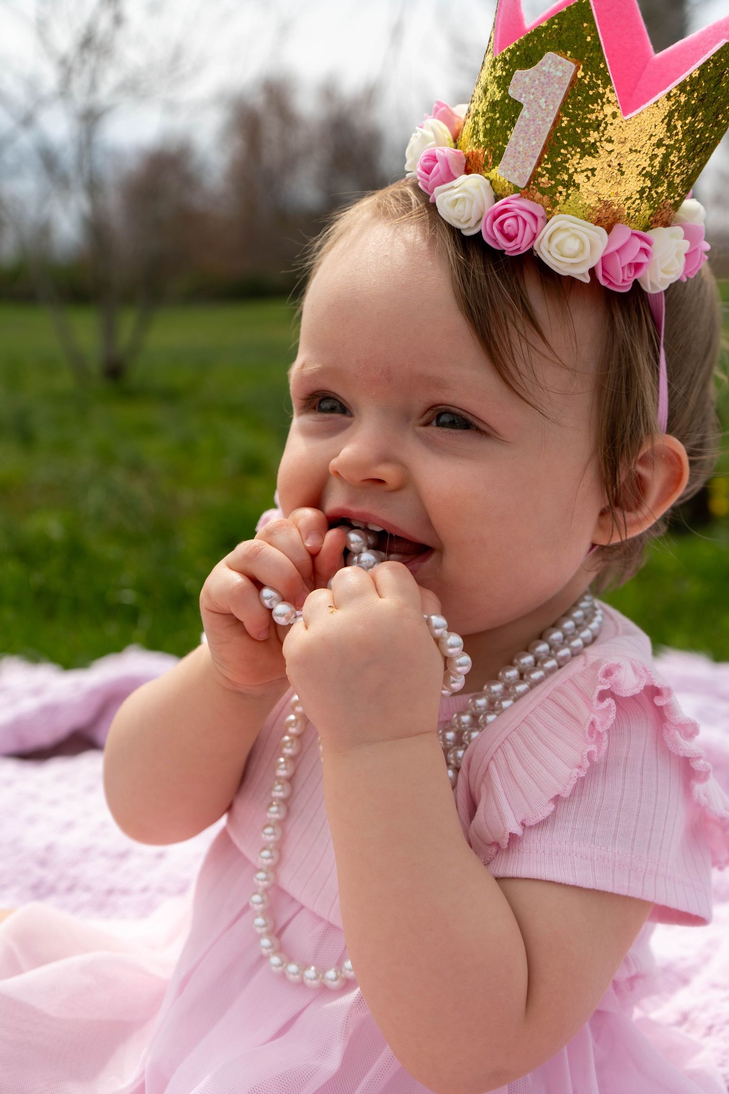 Adorable baby girl in pink dress and number one birthday crown chewing on a pearl necklace outdoors.