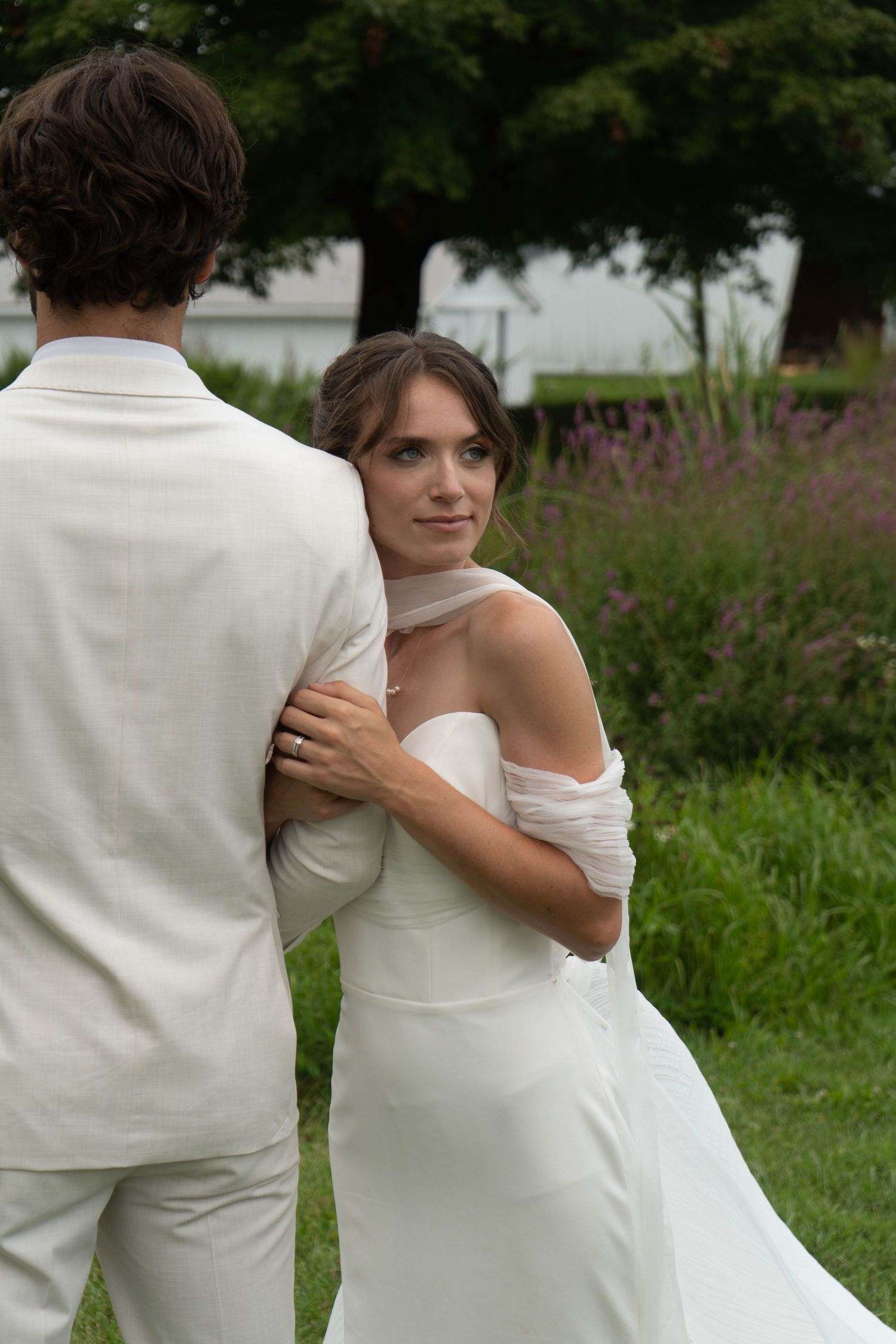 Bride in off-shoulder white gown embraces groom outdoors among purple wildflowers.
