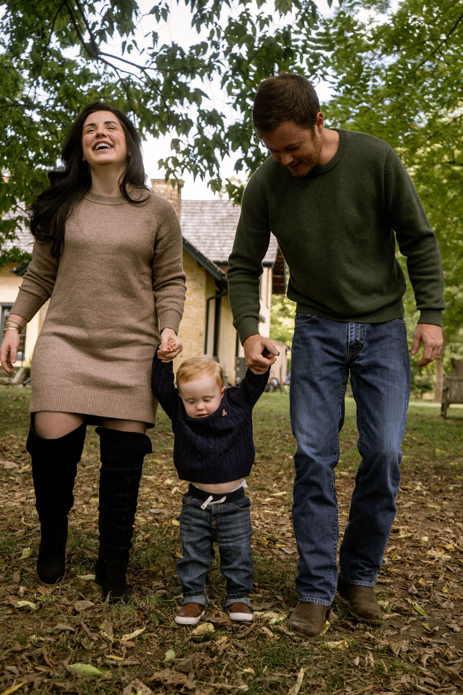 Happy family with mom, dad, and toddler holding hands walking outdoors in a lush green park during fall.