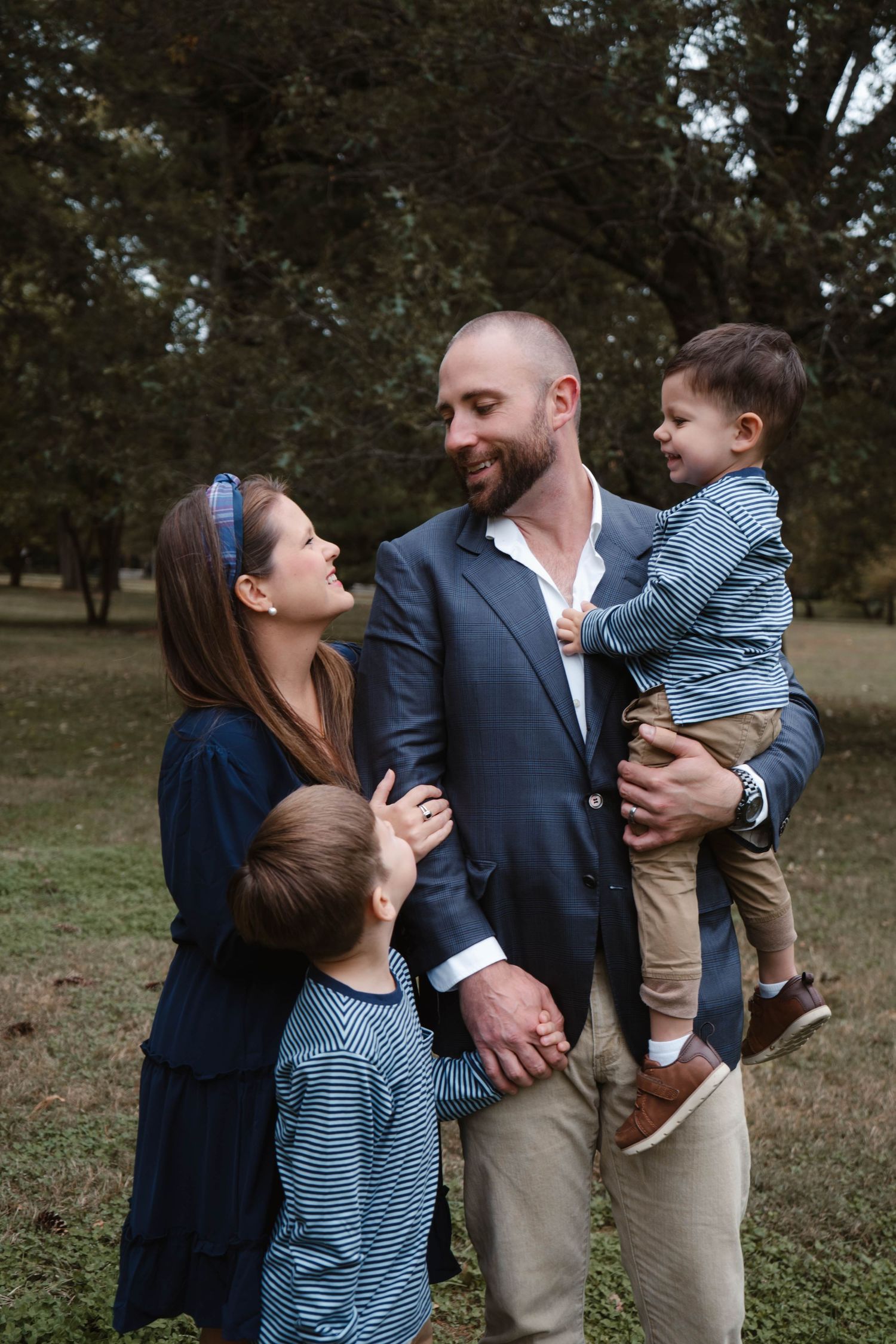 Loving family of four shares a joyful moment outdoors, father holding toddler as mother and older son look up smiling.