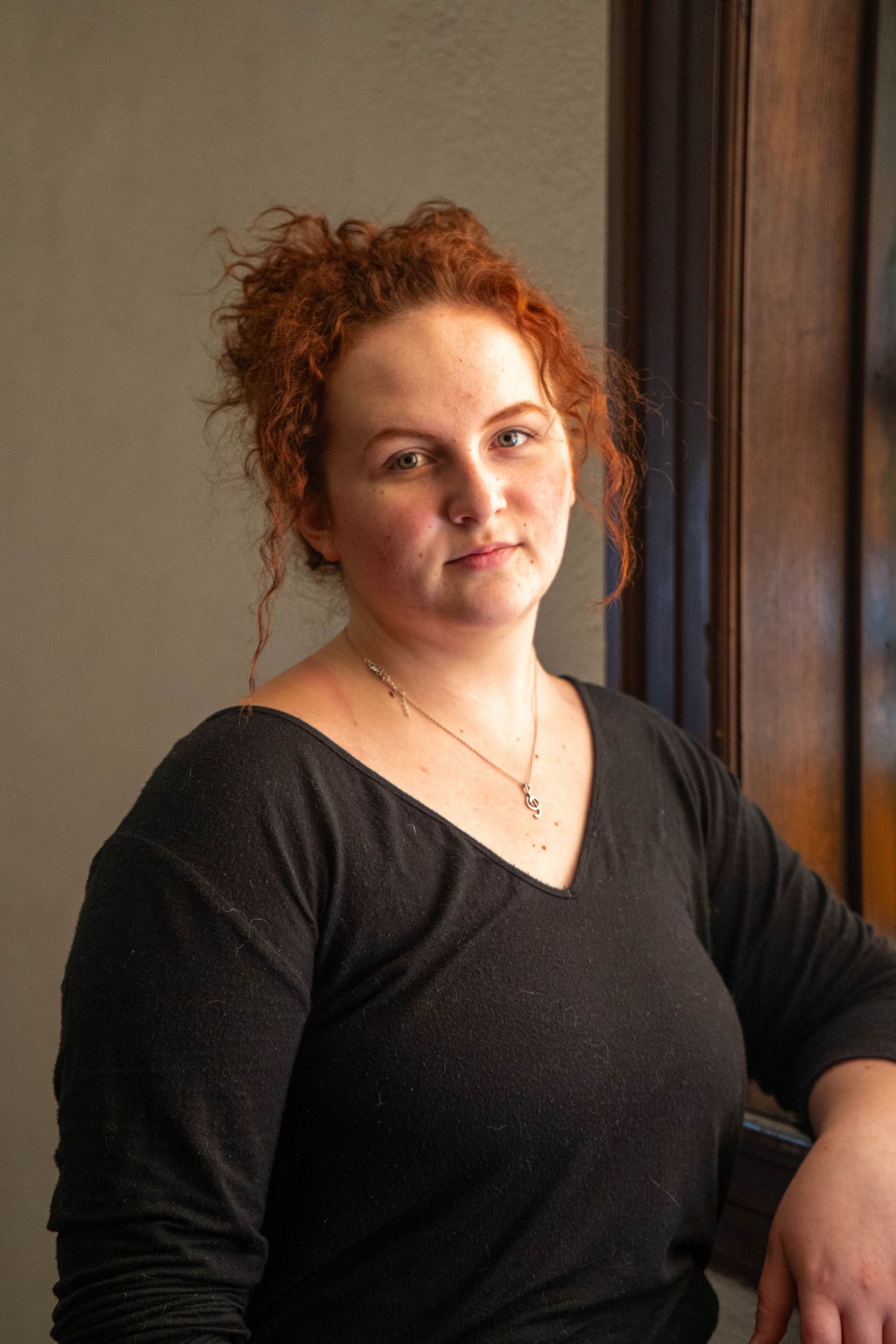 Young woman with curly red hair in black v-neck top leaning against wall in natural light.