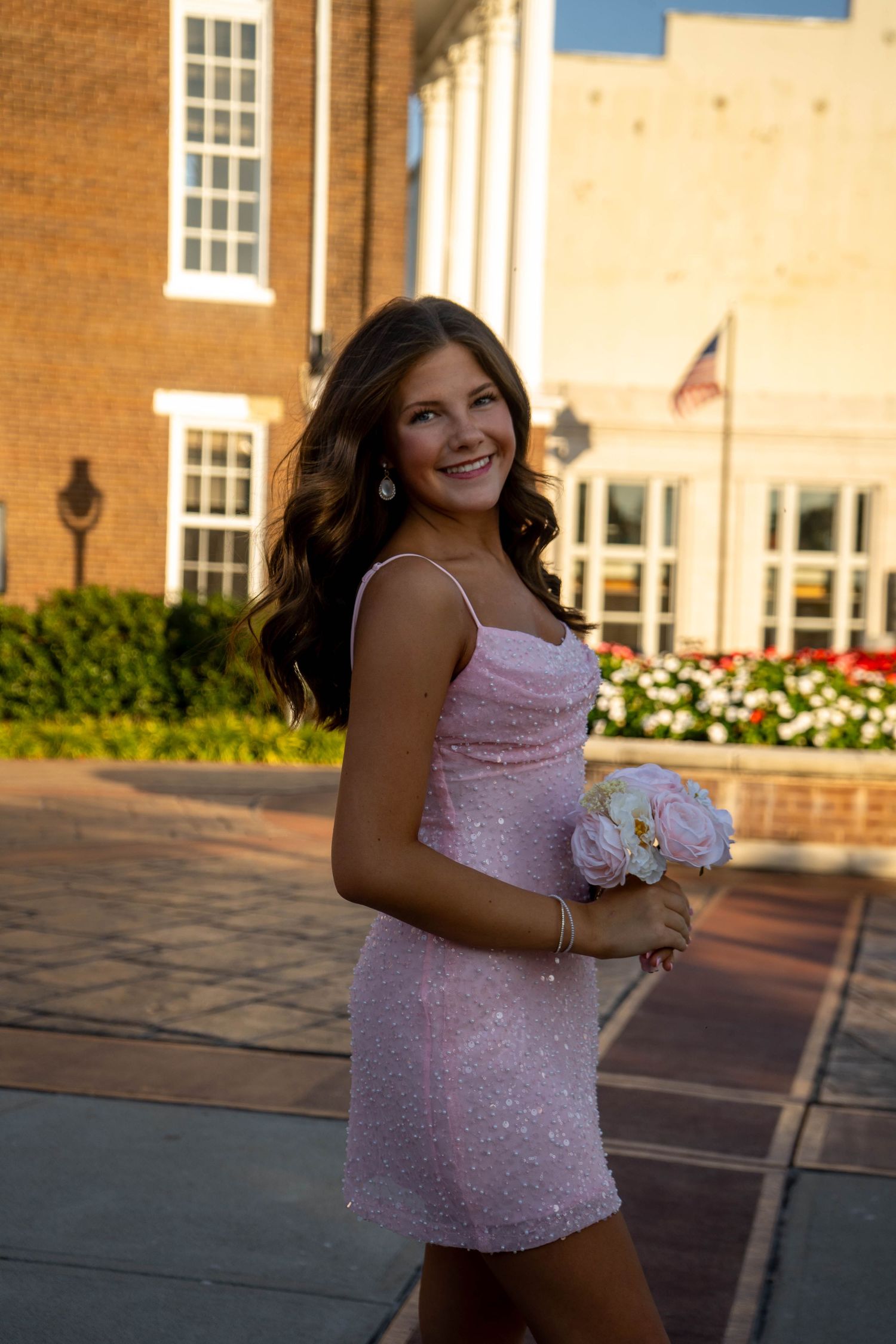A smiling brunette teen in a pink sequin dress holds a white floral bouquet at golden hour.