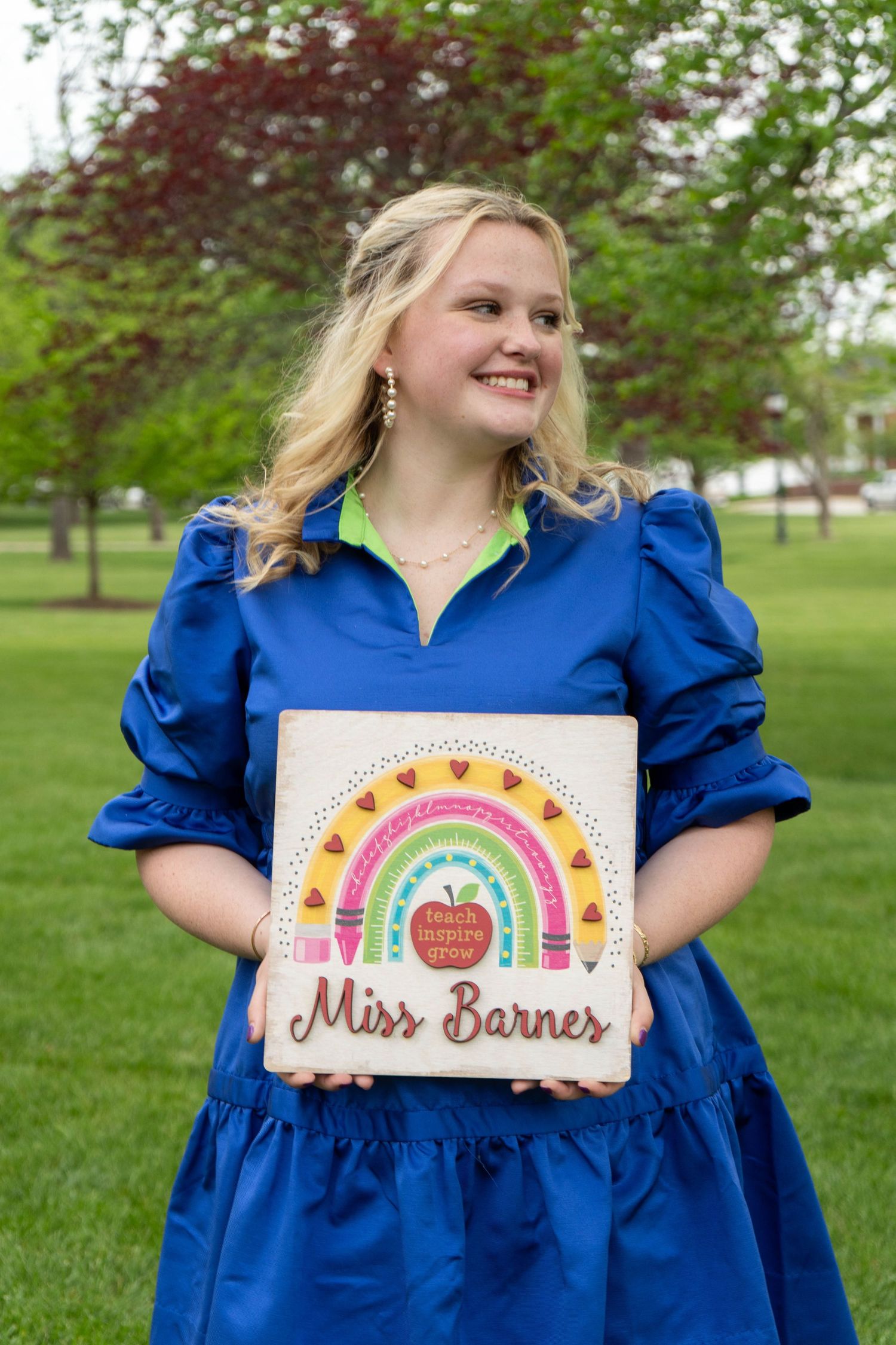 Graduate in blue dress holding Miss Barnes teacher-themed decorative sign outdoors.