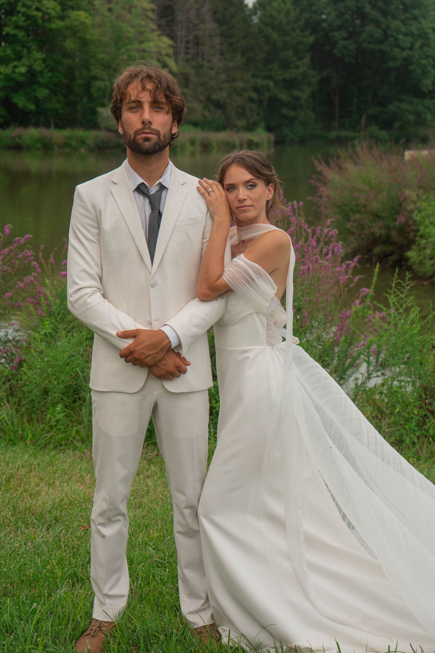 Elegant bride and groom posing outdoors near a lake, wearing white wedding attire surrounded by wildflowers.
