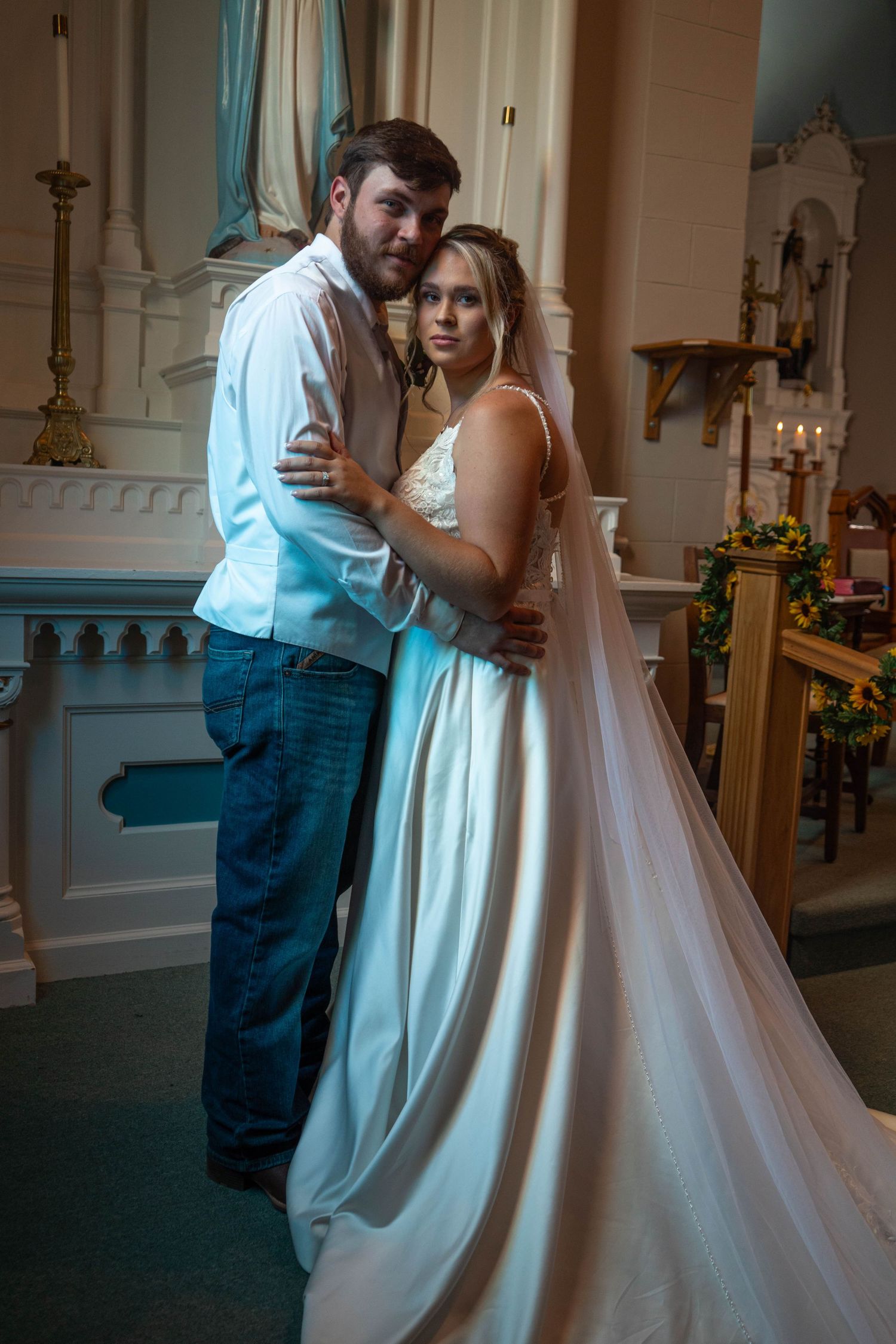 Bride in white gown and groom in casual attire embrace tenderly inside a historic church.