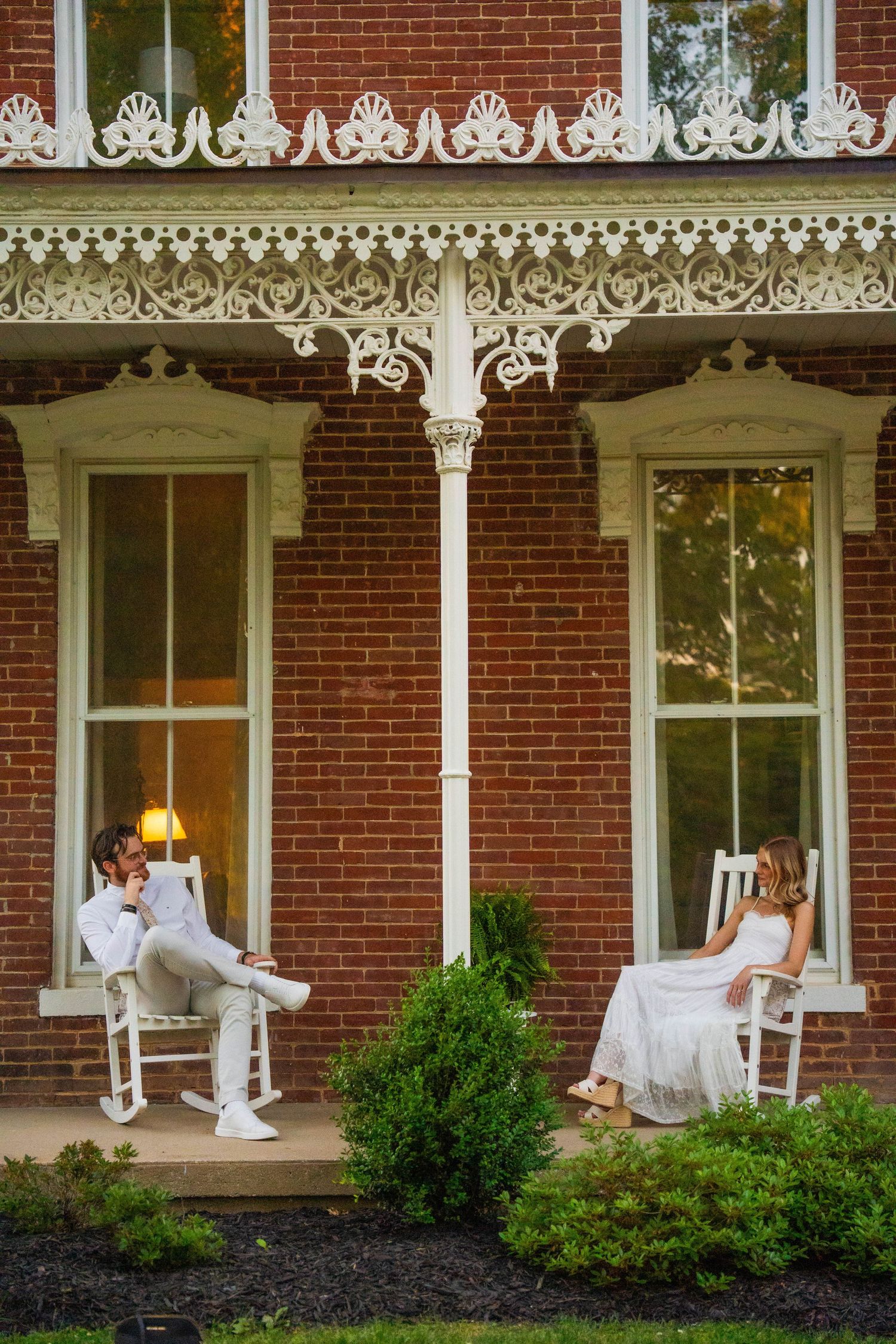 Engaged couple sitting in white rocking chairs on ornate Victorian brick home porch at dusk.