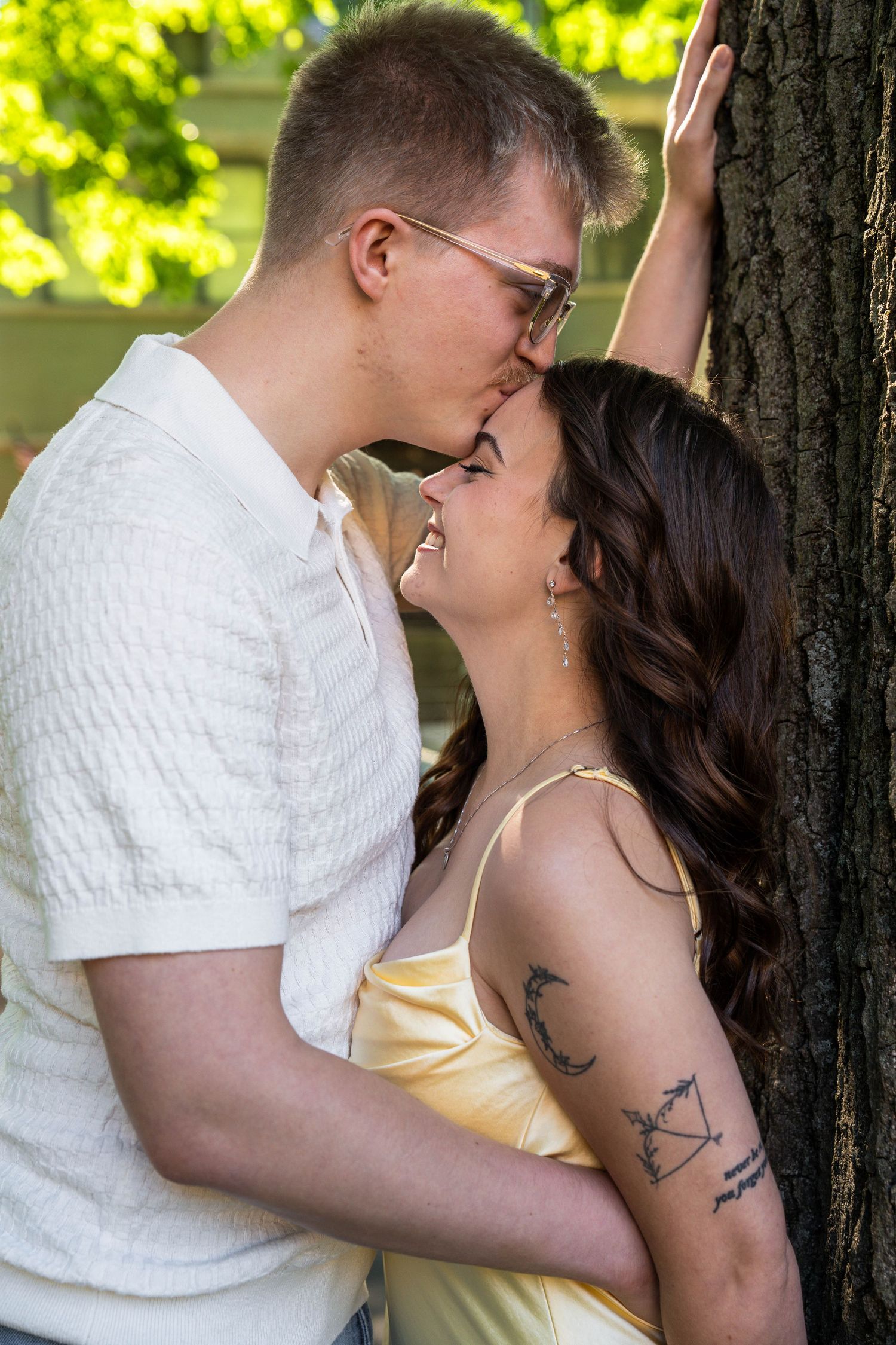 Couple sharing a kiss leaning against a tree in a sunny park setting.