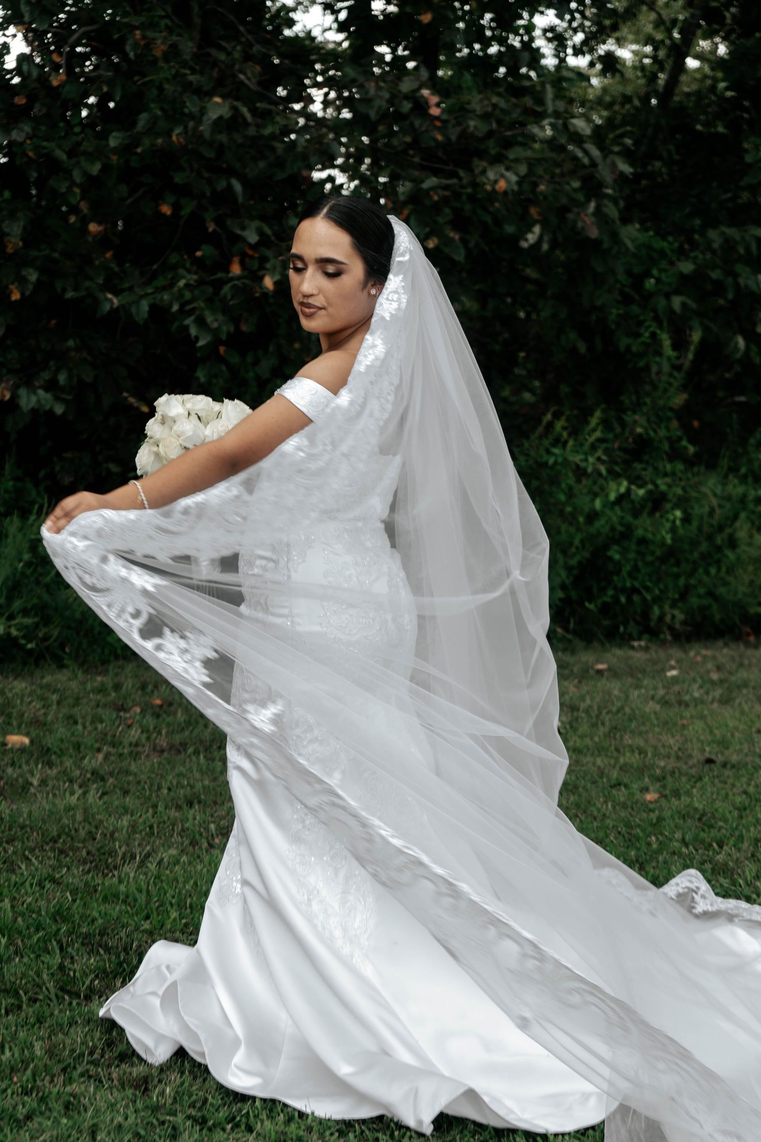 Elegant bride in off-shoulder lace gown and cathedral veil holding white bouquet in garden setting.