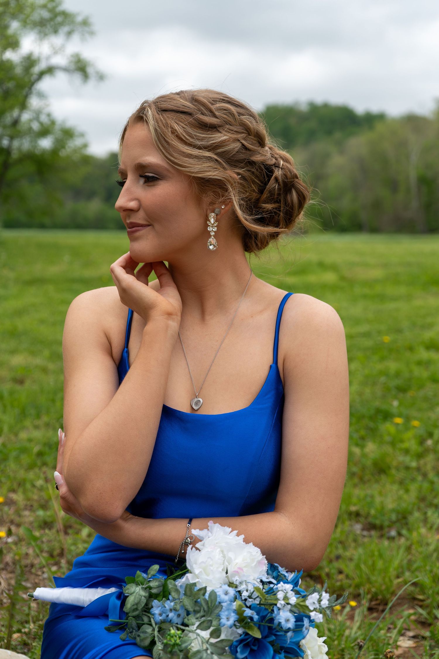 Elegant young woman in royal blue dress sitting in grass, wearing crystal earrings and necklace, holding floral bouquet.