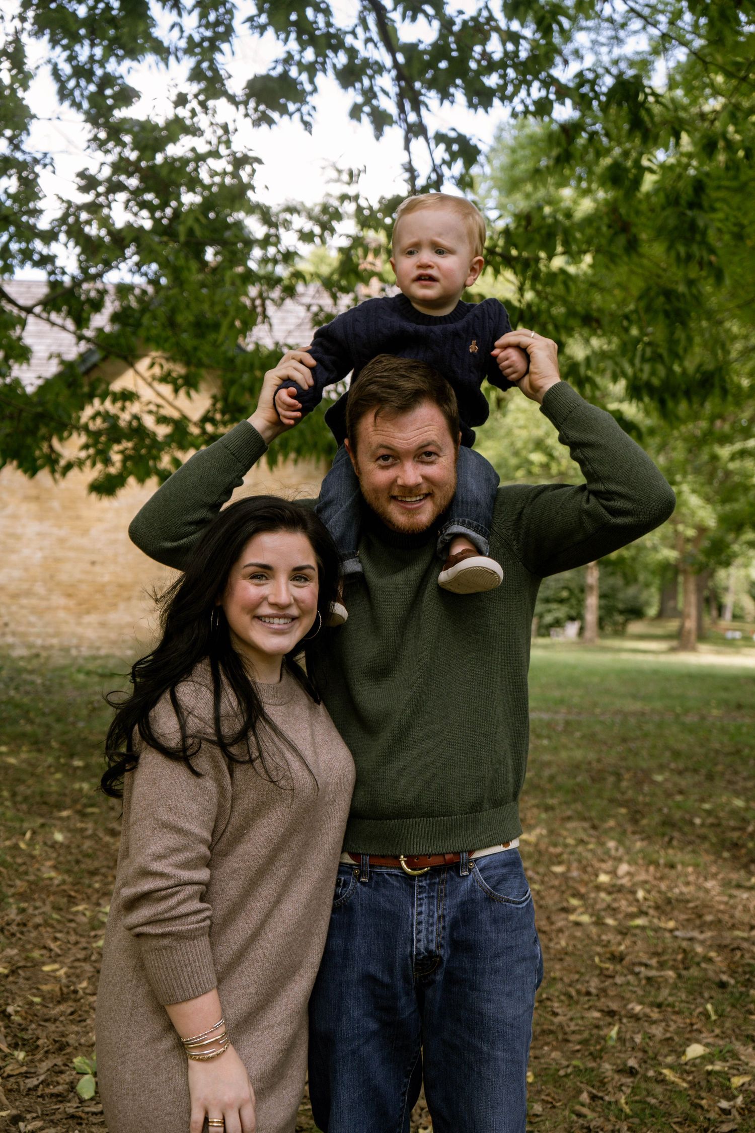 Smiling family portrait with dad holding toddler on shoulders and mom beside them under green trees.